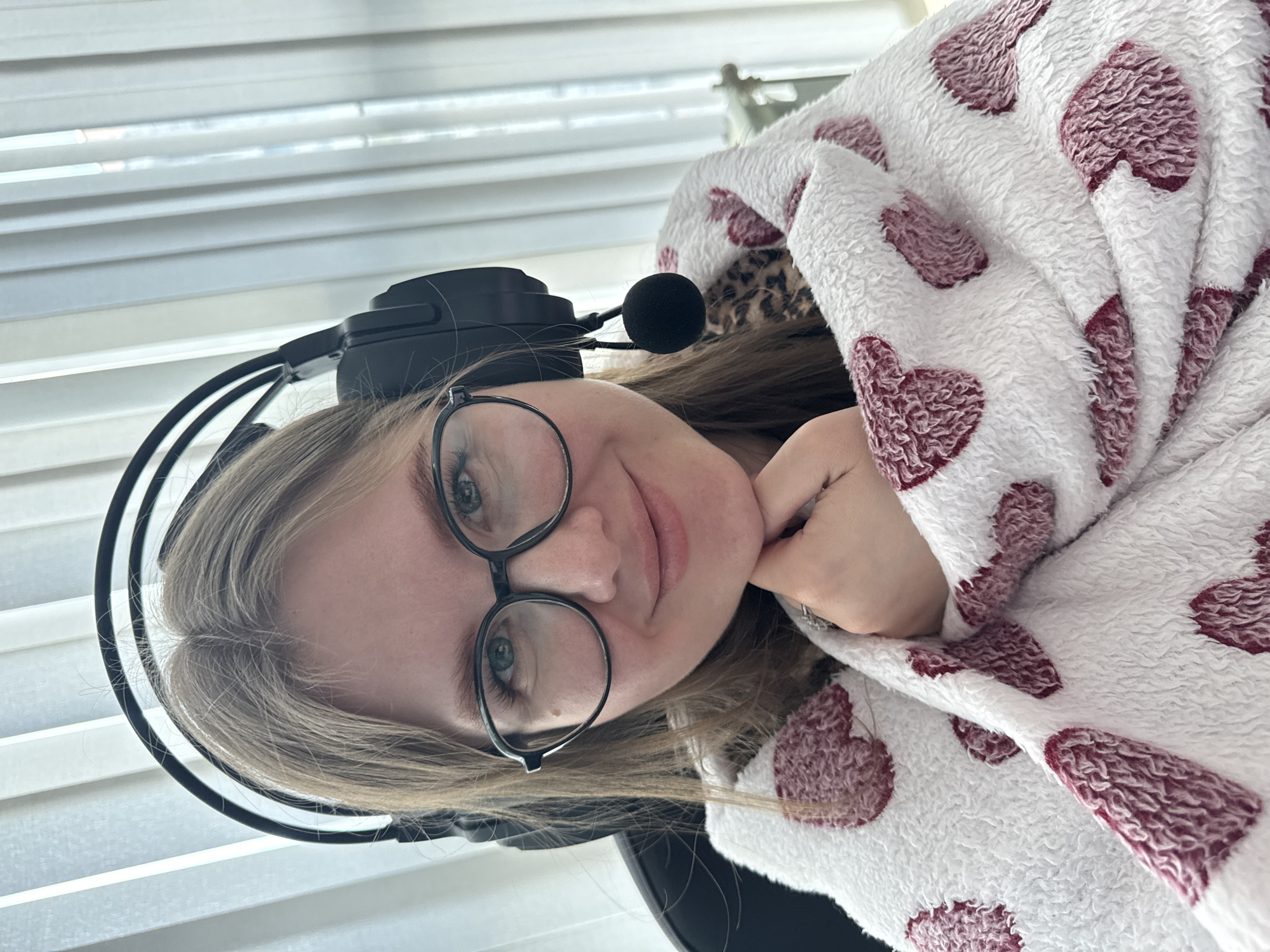 A person with light hair, wearing black-rimmed glasses and a black headset with a microphone, is partially wrapped in a white blanket with maroon heart patterns. The person's hand is visible near their chin. The background shows a window with blinds. The location cannot be confirmed as Perm, Russia from the image provided.