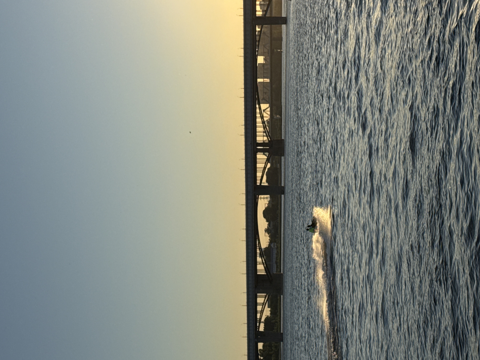 The image captures a serene view of the Ob River in Novosibirsk, Russia, during the late afternoon or early evening. A large, multi-span bridge dominates the horizon, with its concrete piers and roadway clearly visible. The sky transitions from a clear, light blue at the top to a warm, golden hue near the horizon, indicating the setting sun.
In the foreground, a jet ski with two occupants, silhouetted against the bright water, cruises across the river, leaving a trail of white spray. The water is calm, with gentle ripples reflecting the soft light. The riverbank is lined with trees and structures, suggesting a semi-urban or park-like setting on the far side. The overall atmosphere is peaceful and tranquil, highlighting the natural beauty of the river and the impressive engineering of the bridge.