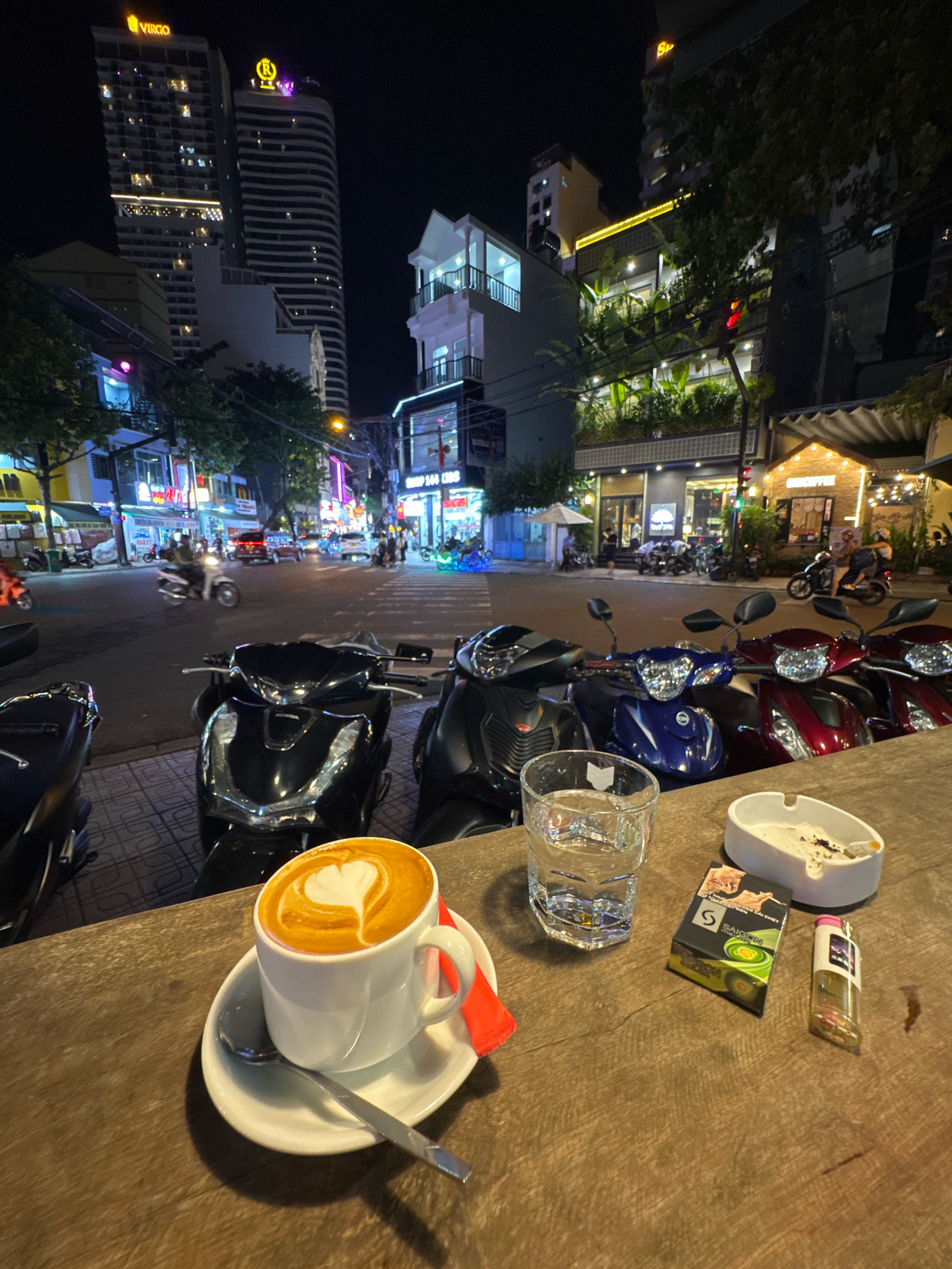 The image depicts a street scene at night in Nha Trang, Vietnam, likely from a cafe or outdoor seating area. In the foreground, a wooden table holds a white cup of coffee with latte art resembling a heart, a glass of water, a pack of cigarettes, a lighter, and an ashtray with cigarette butts. Behind the table, several motorcycles are parked. The background shows a busy city street at night, with traffic including cars and motorcycles, illuminated buildings, and neon signs. Tall buildings with lights are visible in the distance. The overall atmosphere is that of a lively urban night setting, possibly a commercial district in Nha Trang.