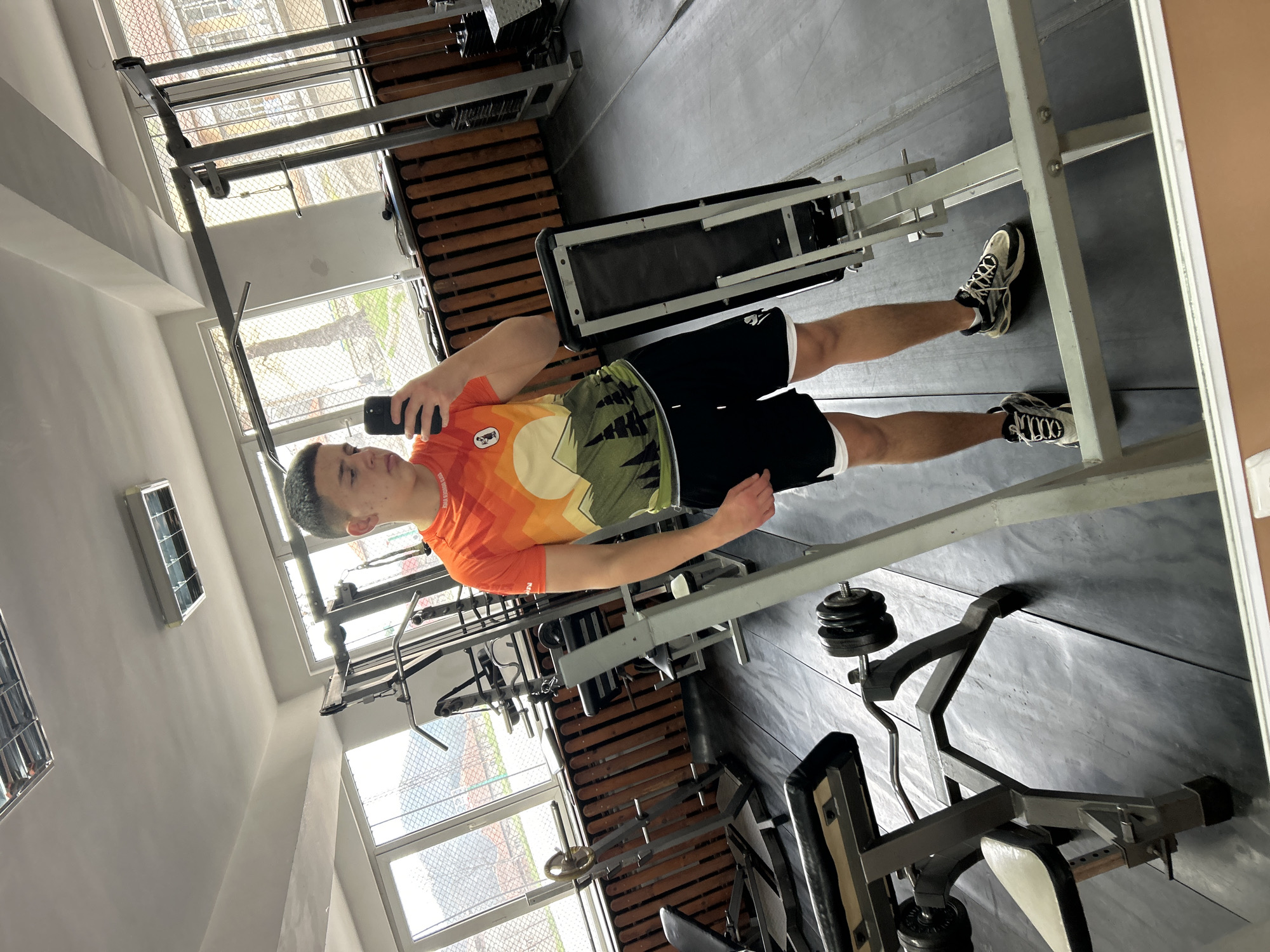 A young man stands in a gym in Zlatograd, Bulgaria, taking a selfie. He is wearing an orange t-shirt with a mountain design, black shorts with white trim, and sneakers. He holds a black smartphone in his left hand. The gym equipment includes various weight machines, a leg press, and a bench. Windows are visible in the background. The walls are painted white.