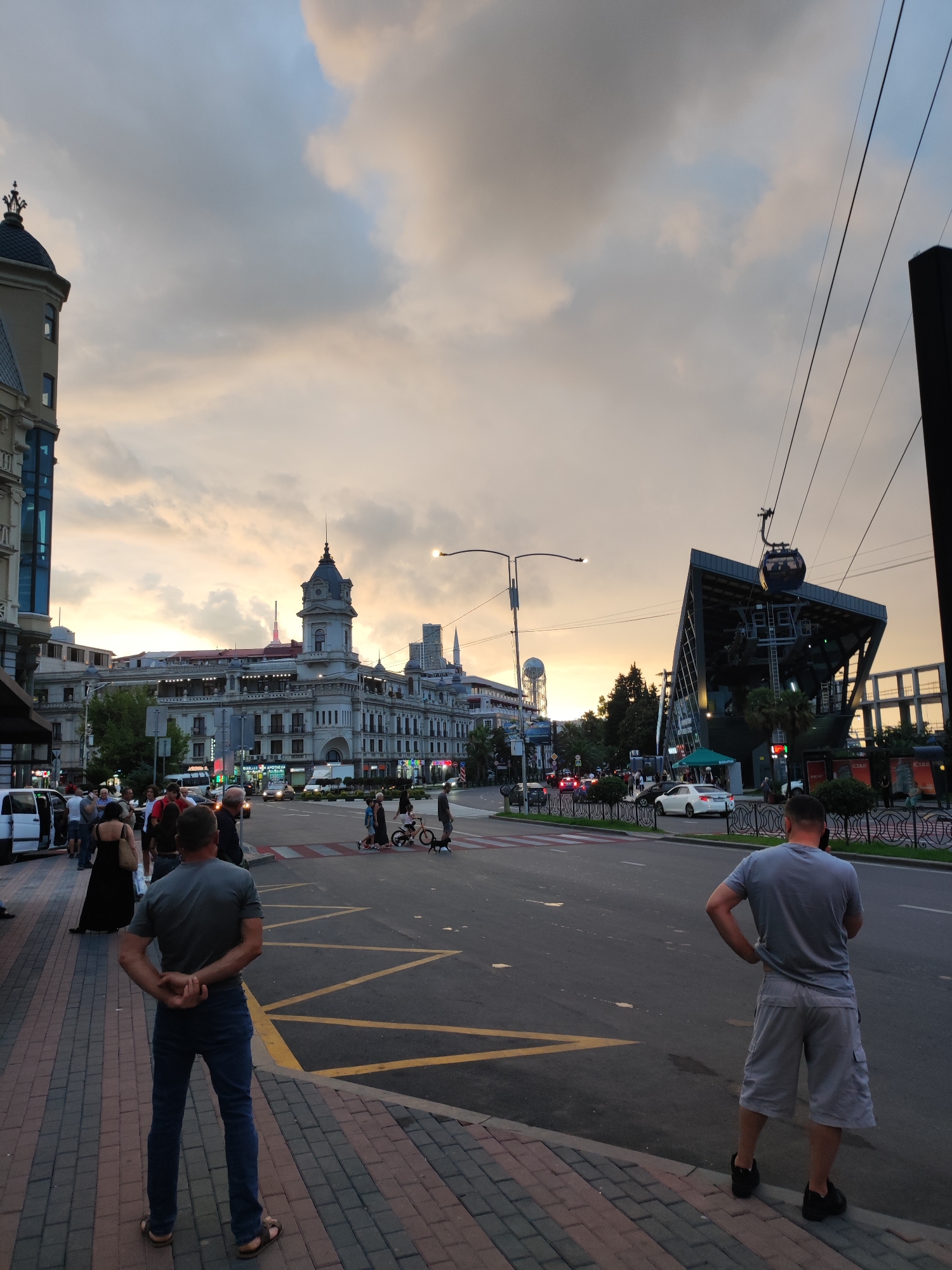 A wide shot captures a street scene at dusk in an urban environment. The sky is filled with dramatic, cloudy formations tinged with sunset colors of orange and yellow. In the foreground, two men stand on a sidewalk, facing away from the camera, with their backs to the viewer. The man on the left wears a grey t-shirt and blue jeans, with his hands clasped behind his back. The man on the right wears a grey t-shirt and light-colored shorts. The sidewalk is paved with brick in alternating shades of red and grey.
The street itself is paved with asphalt and marked with yellow lines. Several cars are visible on the road, some in motion and others stopped. On the left side of the street, a large, ornate building with a distinctive tower and domed roof dominates the background. To the right, a modern cable car station with a sloping roof and supporting cables is prominent. A cable car is suspended in the air, nearing the station. Further in the background, more buildings and trees line the street, and the lights of businesses are beginning to illuminate. A crowd of people are seen walking on the sidewalk on the left, and some are crossing the street. The overall impression is of a busy city street during the transition from day to night.
