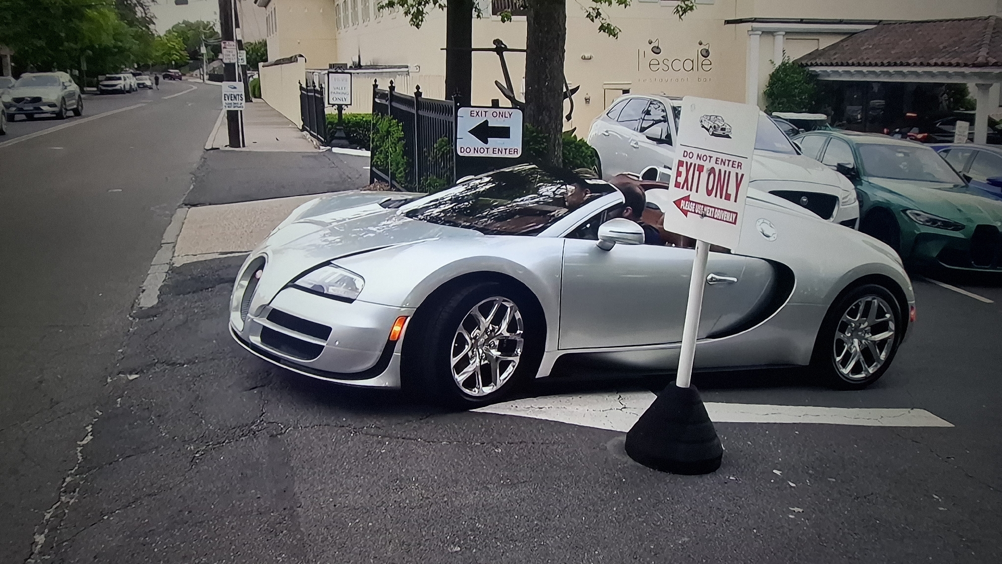 A silver Bugatti Veyron Grand Sport Vitesse convertible is prominently featured in the foreground, appearing to be slowly navigating or exiting a driveway on an asphalt surface. Two male occupants are visible inside the car, with the driver wearing a hat or cap.
The setting suggests an upscale location, possibly a private entrance or valet area for a restaurant or event. A light-colored building with a tiled roof is visible in the background, featuring a sign that reads "l'escale restaurant bar". Black ornate fencing separates the street from the property.
Several signs are present, indicating a specific traffic flow. On a street pole to the left, signs read "EVENTS" and "EXIT ONLY" with a left-pointing arrow, along with "DO NOT ENTER". In the immediate foreground, a portable stand-up sign features a diagram of a car and clearly states "DO NOT ENTER EXIT ONLY" with a large red arrow pointing left, adding the instruction "PLEASE USE NEXT DRIVEWAY". Another partially visible sign behind the fence reads "TOYS VALET PARKING ONLY".
Other vehicles are parked or visible in the background, including a white SUV on the left street, and a white sedan/SUV (possibly a Jaguar) behind the Bugatti, with a distinctive green BMW M3/M4 (G80/G82 generation) further back on the right.
The scene takes place during the daytime, under what appears to be an overcast sky, providing soft, diffused lighting. The road surface shows some cracks and wear.