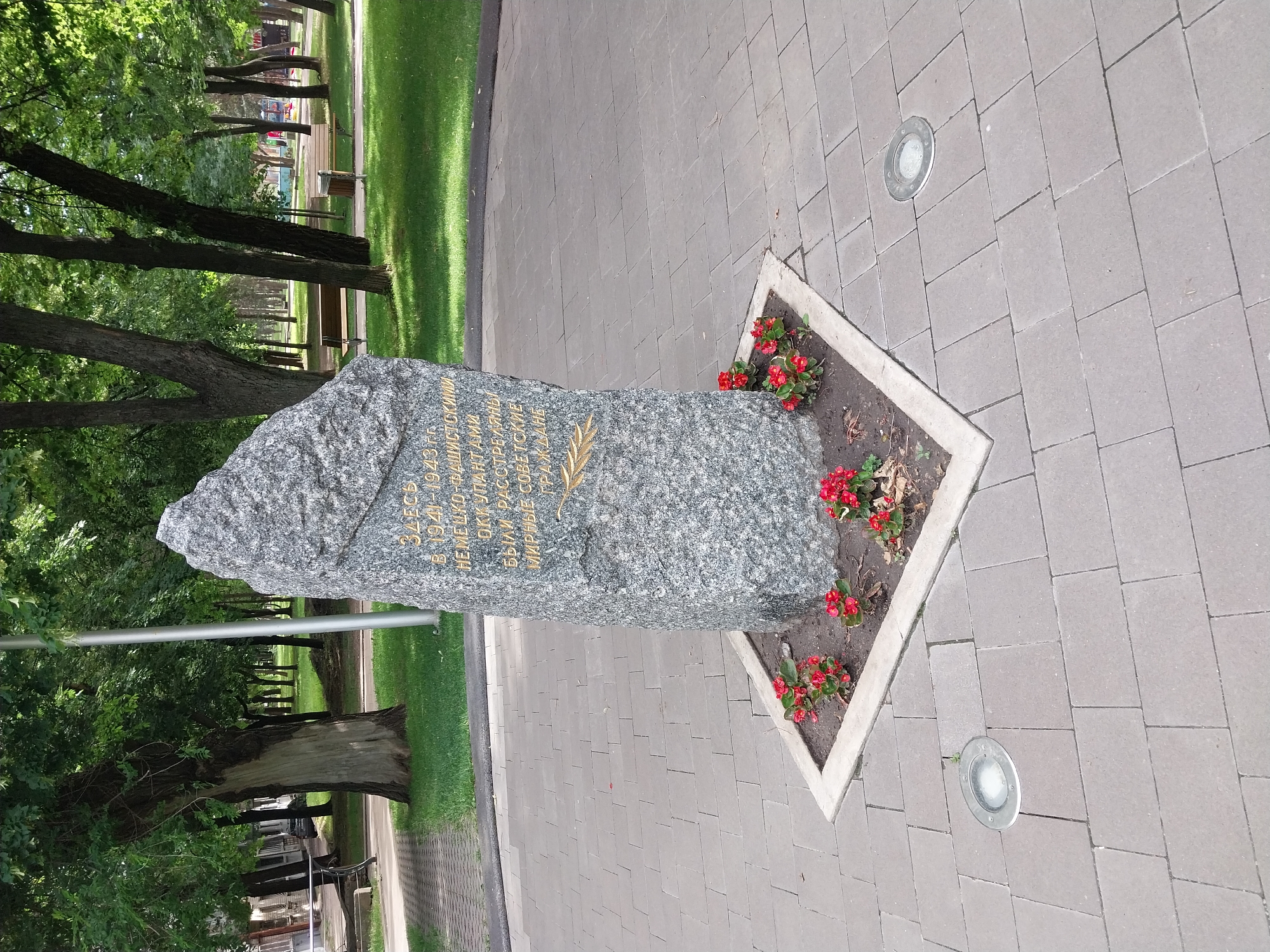 This is a medium, eye-level shot of a memorial monument in a park, located in the city of Dnipro, Ukraine. The monument is a rough, grey stone slab with golden text inscribed on it, and a golden wheat stalk image. The text appears to be in Cyrillic script. The monument is surrounded by a small bed of red flowers and enclosed by a concrete border. The monument stands on a paved area made of grey rectangular bricks. Two circular, flush-mounted lights are visible in the paving. In the background, there are several trees with green foliage, grassy areas, park benches, and a children's playground structure. The overall environment suggests a well-maintained public park in an urban setting. There are no people visible in the frame.