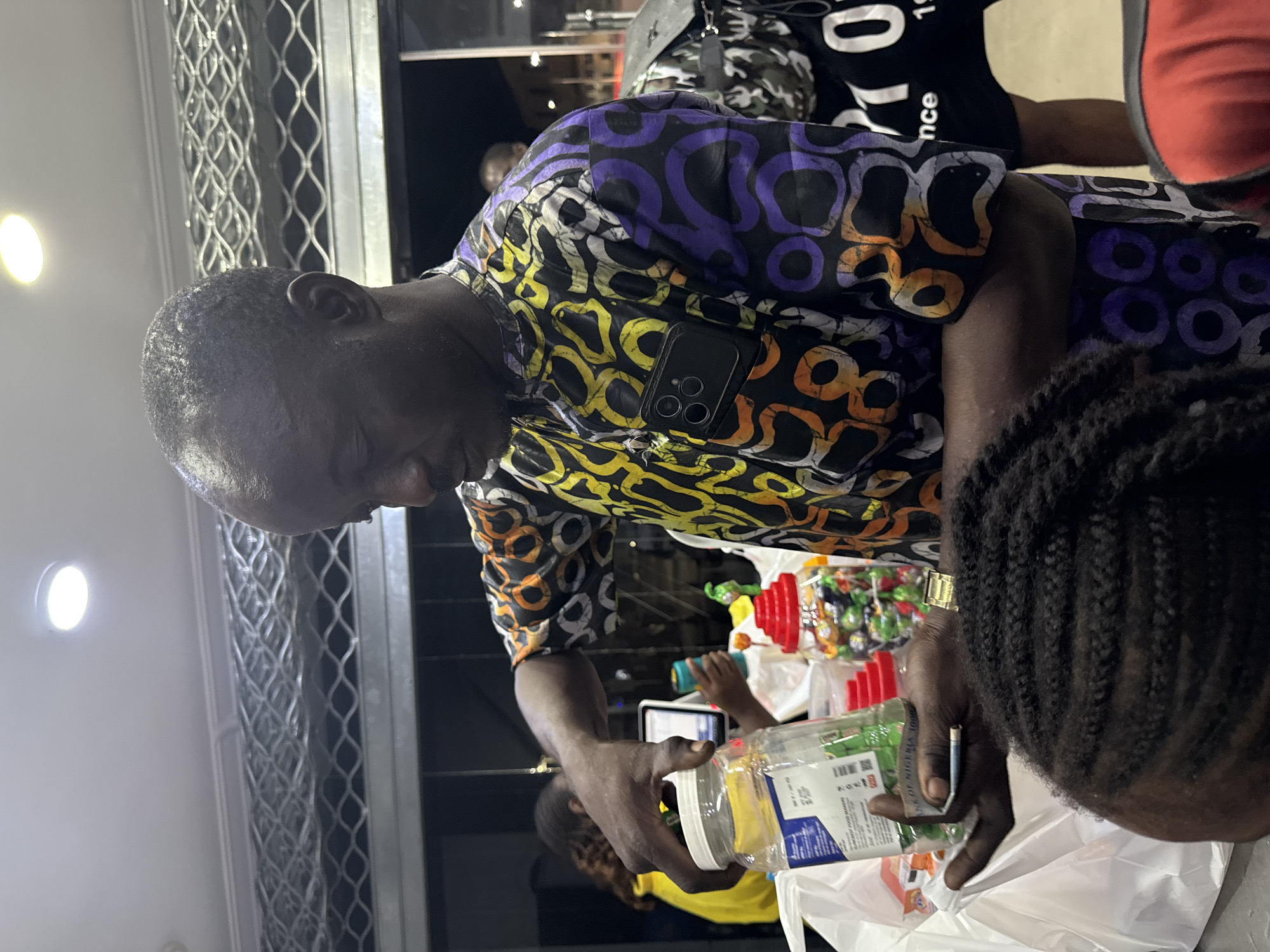 This indoor scene is located in the city of Ado, Nigeria.
An adult Black male is centrally positioned, holding a clear plastic jar with a white screw-top lid in his right hand, while his left hand is near the bottom of the jar, also grasping a small, thin, cylindrical object. He is looking down at the jar or its contents. He wears a short-sleeved, collared shirt patterned with abstract circular and ring shapes in yellow, orange, purple, white, and black. A dark-cased smartphone with a multi-lens camera array is tucked into his left chest pocket. A gold-colored watch is visible on his left wrist. The jar contains green-wrapped items, likely candies, and features a white label with blue and black text, including "SUPERIOR FOOD MAKERS" and a partial reference to "BANK OF NIGERIA 1000".
The environment appears to be a shop or market. Above the man, a white ceiling with recessed circular LED lights is visible, along with a metal mesh structure. To the man's right, on a counter, are several other clear plastic jars with red lids, filled with various colorful sweets. A white plastic shopping bag is present near his left hand. In the out-of-focus background to the left, a person with reddish-brown hair wearing a yellow top is partially visible, seemingly interacting with a tablet device. To the right, another person in a black t-shirt with white lettering is partially obscured. In the immediate foreground, at the bottom center, the top of a child's head with dark braided hair is visible.