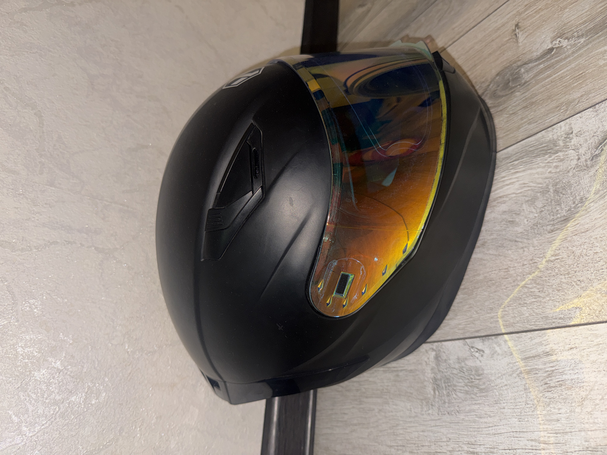 A black motorcycle helmet with a reflective orange-tinted visor rests on a light-colored wooden floor in front of a textured, light-colored wall. The helmet is positioned slightly angled towards the right. The visor shows streaks and smudges. A dark horizontal bar is visible on the wall behind the helmet. The overall lighting is dim. The scene appears to be indoors.