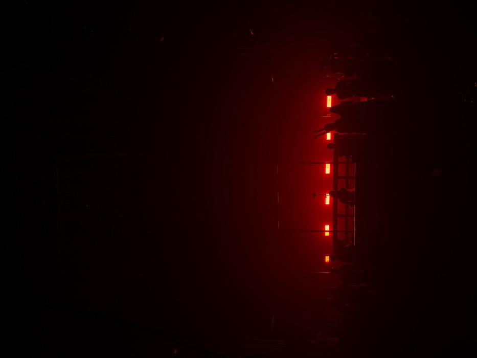 A dark, blurry image is illuminated by a series of stacked, rectangular red lights. Silhouettes of figures surround the light source, obscuring some of the glowing shapes. The scene suggests an urban setting, likely at night, with potential construction or emergency lighting present. The overall impression is one of mystery and implied danger.