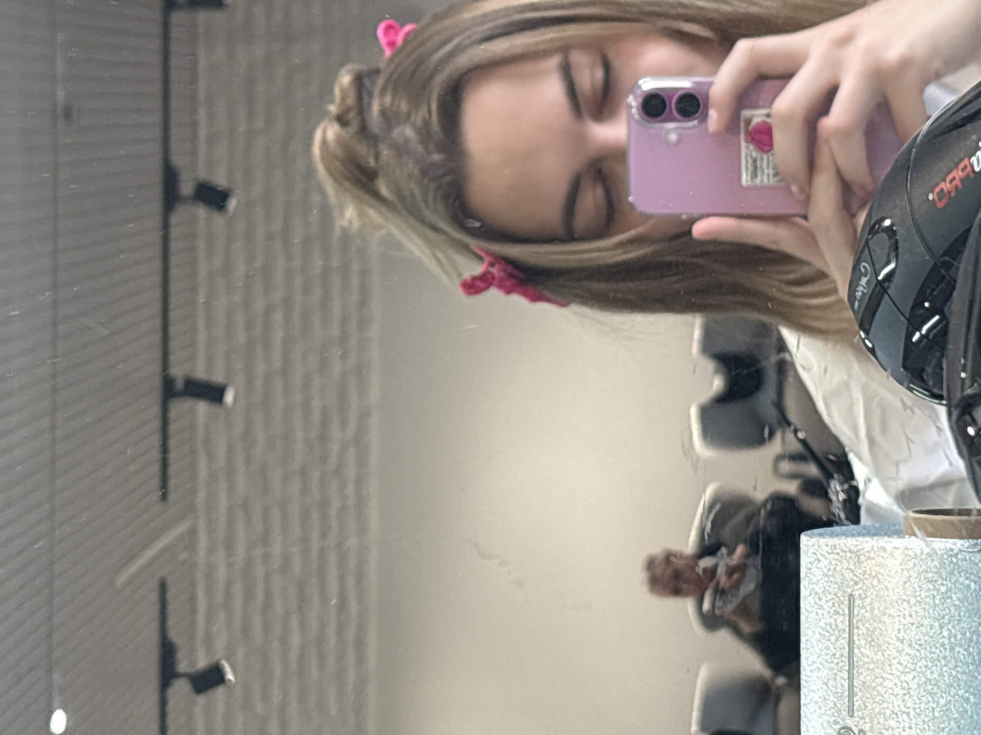 A mirror selfie shows a young woman with blonde hair secured by pink clips. She is holding a pink cell phone, presumably taking the photo, in front of her closed eyes. Reflected in the mirror is a salon setting, including salon chairs, a stylist, and a hairstyling helmet. The exact location in Krasnoyarsk, Russia cannot be determined from the image.