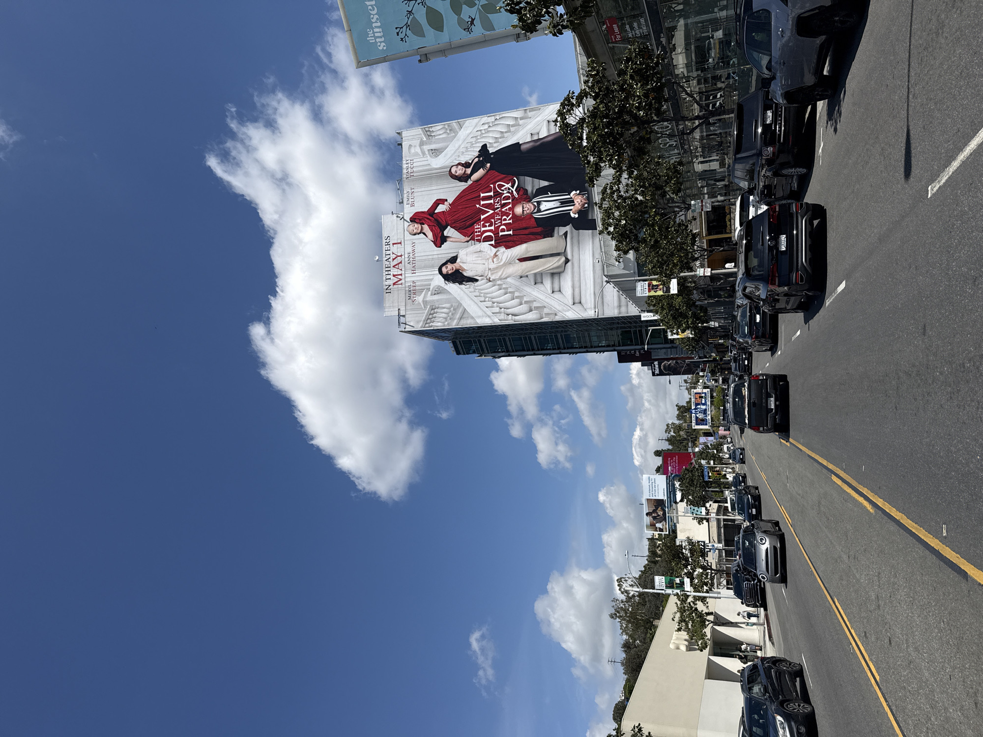 The image depicts a street scene in West Hollywood, United States. Prominently featured is a large billboard advertising "The Devil Wears Prada," with the release date listed as May 1. The billboard displays the names of the actors Meryl Streep, Anne Hathaway, Emily Blunt, and Stanley Tucci. The sky is visible above, showing a mix of blue and puffy white clouds.
The street is lined with parked cars and features double yellow lines. Other billboards and signage are visible further down the road. Trees line the sidewalk. The day appears to be sunny.