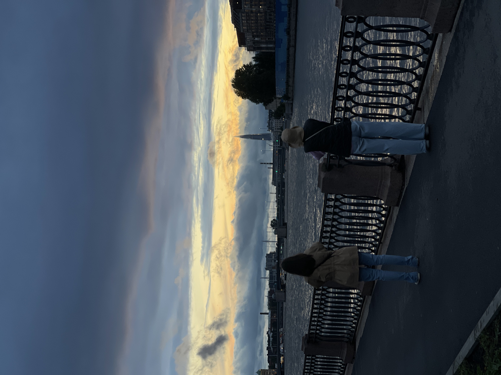 Two individuals stand facing a body of water, observing a distant cityscape under a dramatic twilight sky. The sky displays a gradient of dark blues at the top, transitioning to shades of gray, pink, and yellow closer to the horizon, indicating either sunset or sunrise. A prominent, needle-like skyscraper is visible in the distance, piercing through the cloud cover. The water is choppy, reflecting the muted colors of the sky. A decorative black metal fence separates the walkway from the water's edge. The walkway is paved and appears to be a promenade or embankment. The individuals are dressed casually, one in a light-colored jacket and blue jeans, the other in a darker vest over a light top and blue jeans. The scene is likely Saint Petersburg, Russia, given the architectural style of the distant buildings and the presence of a major river or canal, potentially the Neva River, and the distinct skyscraper which resembles the Lakhta Center.