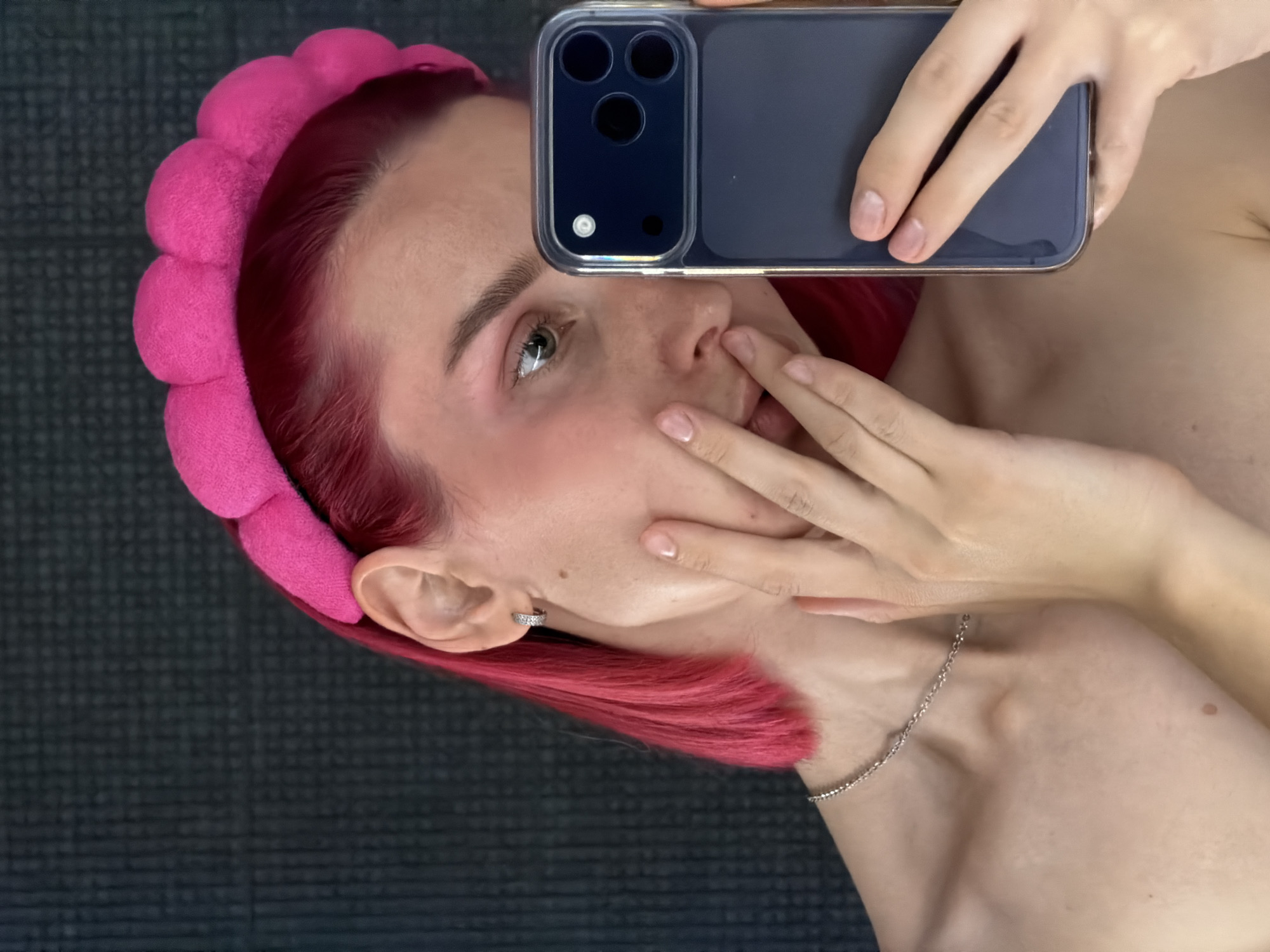 A young woman with bright pink hair and a pink headband is taking a selfie in what appears to be a home environment. She is wearing a silver chain necklace and a small silver hoop earring. Her left hand is resting on her face, while her right hand holds a dark-colored smartphone with a multi-lens camera array. The background is a dark, textured surface, possibly a wall or fabric. There are no visible indications of the city of Chaikovsky, Russia, in the image.