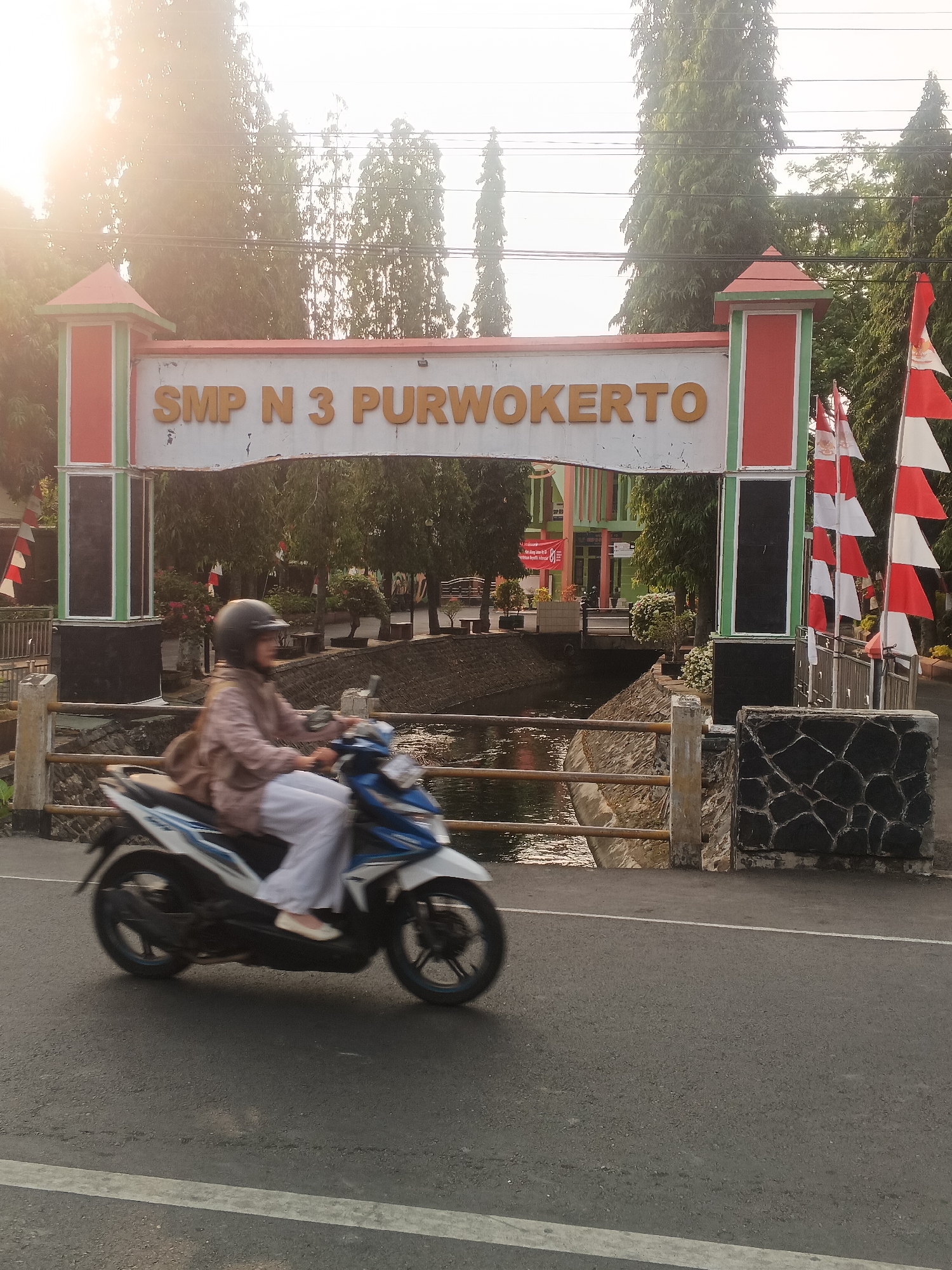The image depicts the entrance to SMP N 3 PURWOKERTO, located in Sokanegara, Indonesia. A person wearing a helmet, light brown jacket, and white pants is riding a white and blue motorcycle from left to right across the frame. The motorcycle is in motion, creating a blur effect. The entrance archway features the school's name in large, orange letters. Red and white Indonesian flags are displayed on the right side of the entrance. Behind the entrance, trees and buildings are visible. A canal or body of water runs beneath a bridge in front of the school. The scene is illuminated by the sun, which appears to be setting or rising, casting a warm glow.