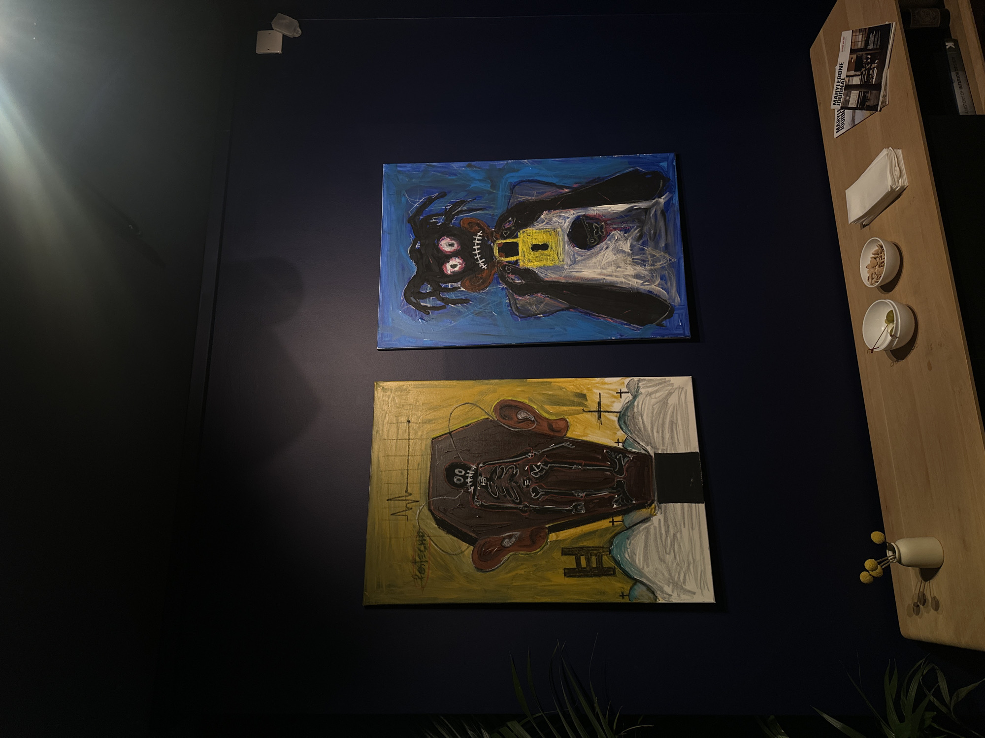 The image depicts a section of a wall with two paintings and a wooden shelf, in an indoor location. The wall is painted a dark blue. The top painting is blue and features a figure with a distorted face and what appears to be a yellow padlock on its chest. The bottom painting is yellow and shows a figure in a coffin. To the right of the paintings, attached to the wall, is a wooden shelf with a light wooden finish. On the shelf, there is a stack of napkins, a small bowl of nuts, a small bowl with what appears to be an olive and a toothpick, a magazine with the words "MASTERS OF INTERIOR" on the cover, and two books. The location is not explicitly stated.