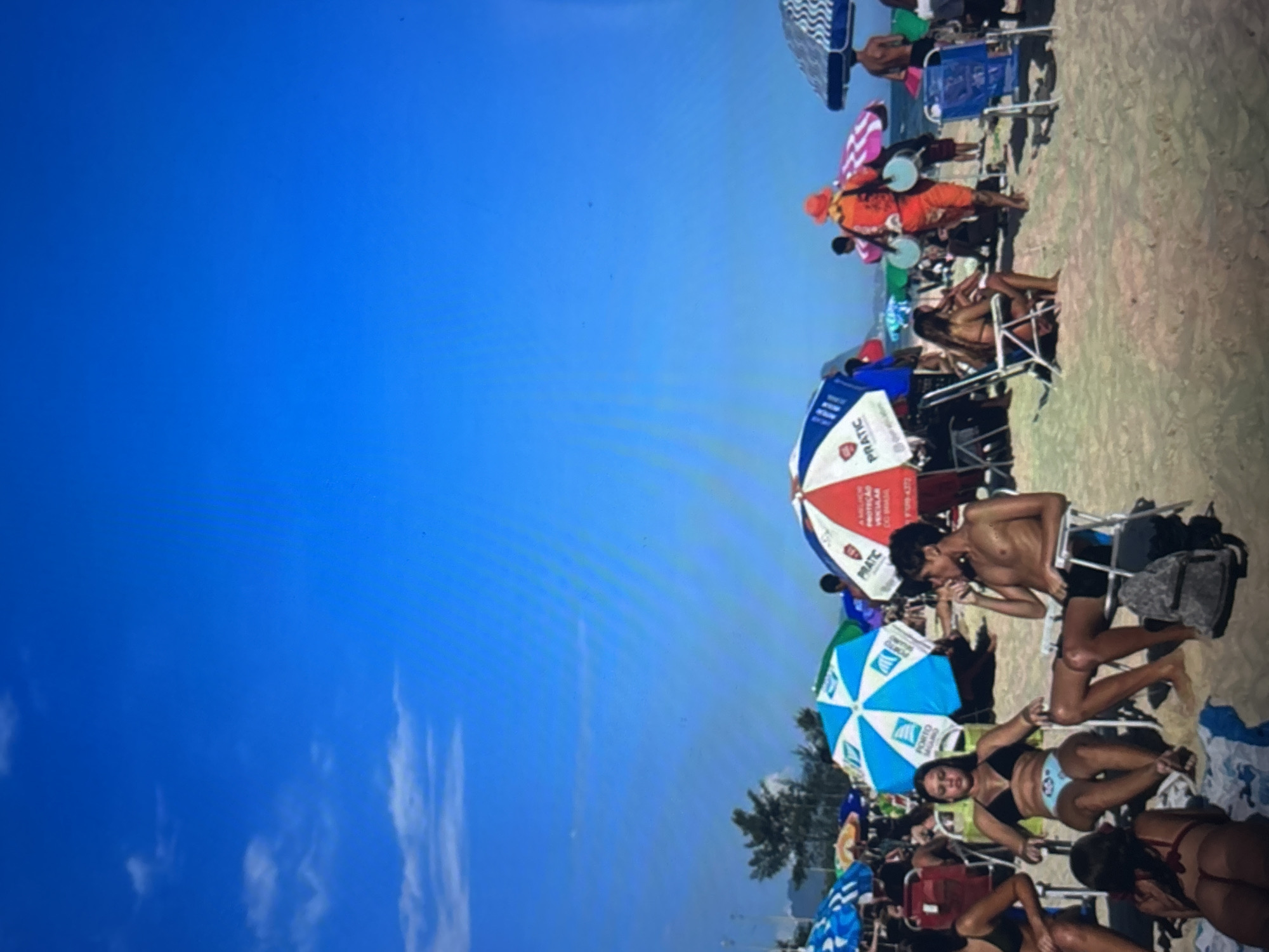 An eye-level, full shot captures a sunny beach scene with many people enjoying the day under umbrellas. In the foreground, a young woman in a black bikini and light blue bottoms sits in a beach chair, looking forward. Next to her, a young man with dark hair and no shirt sits in a beach chair, wearing black swim trunks and holding something to his mouth. Behind them, a woman in a red bikini reclines on a towel.
The beach is populated with numerous umbrellas, some providing shade to beachgoers. A vendor wearing an orange hat and outfit, carrying two light blue cylindrical objects, walks through the scene. People are scattered across the sand, some seated in chairs, others standing or walking. The ocean is not visible, but the bright blue sky suggests a clear day. The background shows distant trees and a mountain range. The sand is light brown and appears well-trodden.