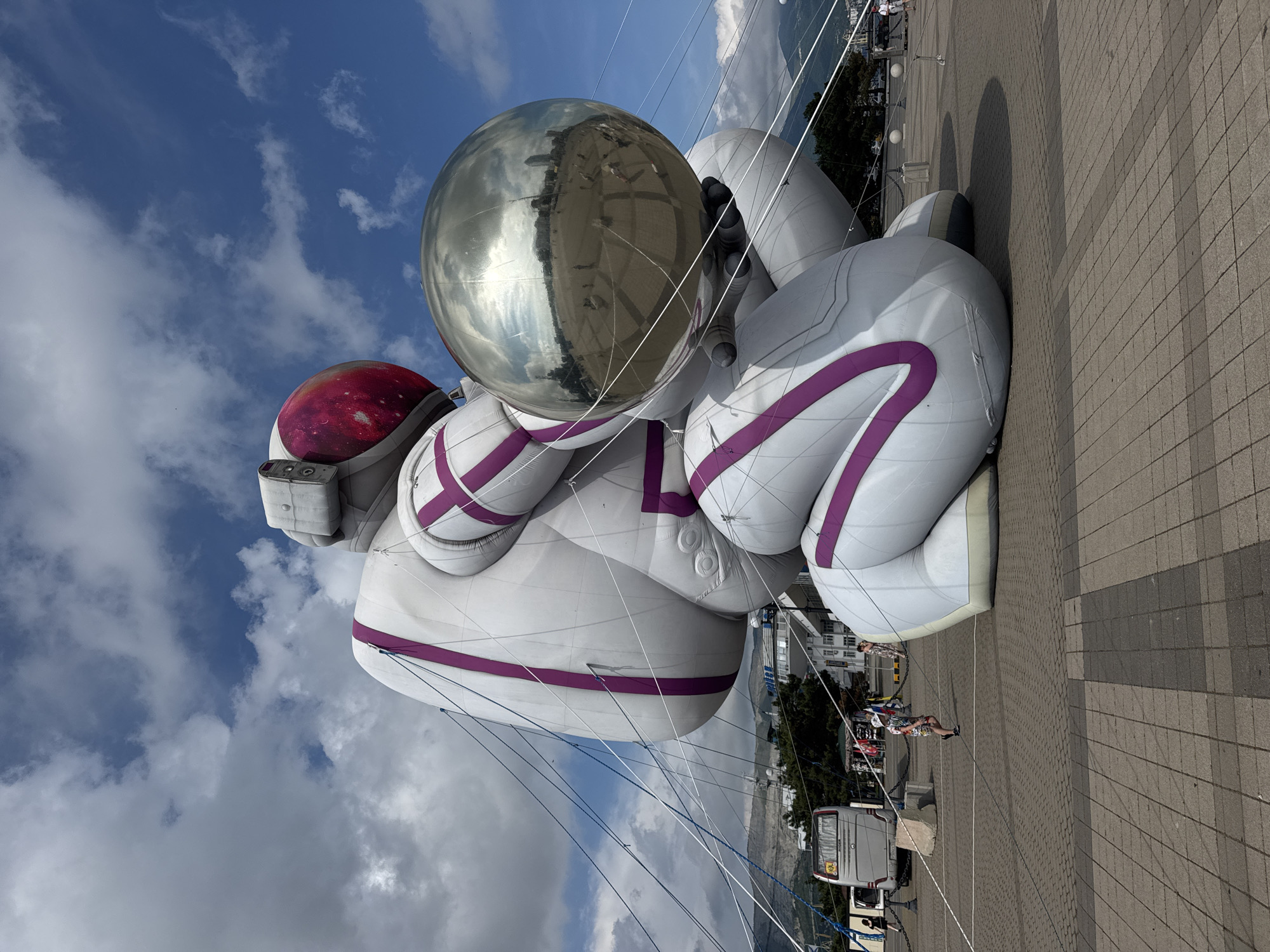 This image depicts a large inflatable sculpture of an astronaut holding a reflective sphere. The sculpture is white with purple accents and appears to be tethered by numerous ropes. In the background, a bus and other vehicles are visible, along with buildings and mountains. Several people are present in the scene, some walking and others appearing to be onlookers. The ground is paved with square tiles. The sculpture is located in Novorossiysk, Russia, specifically at the Embankment of Admiral Serebryakov, in front of the Black Sea.