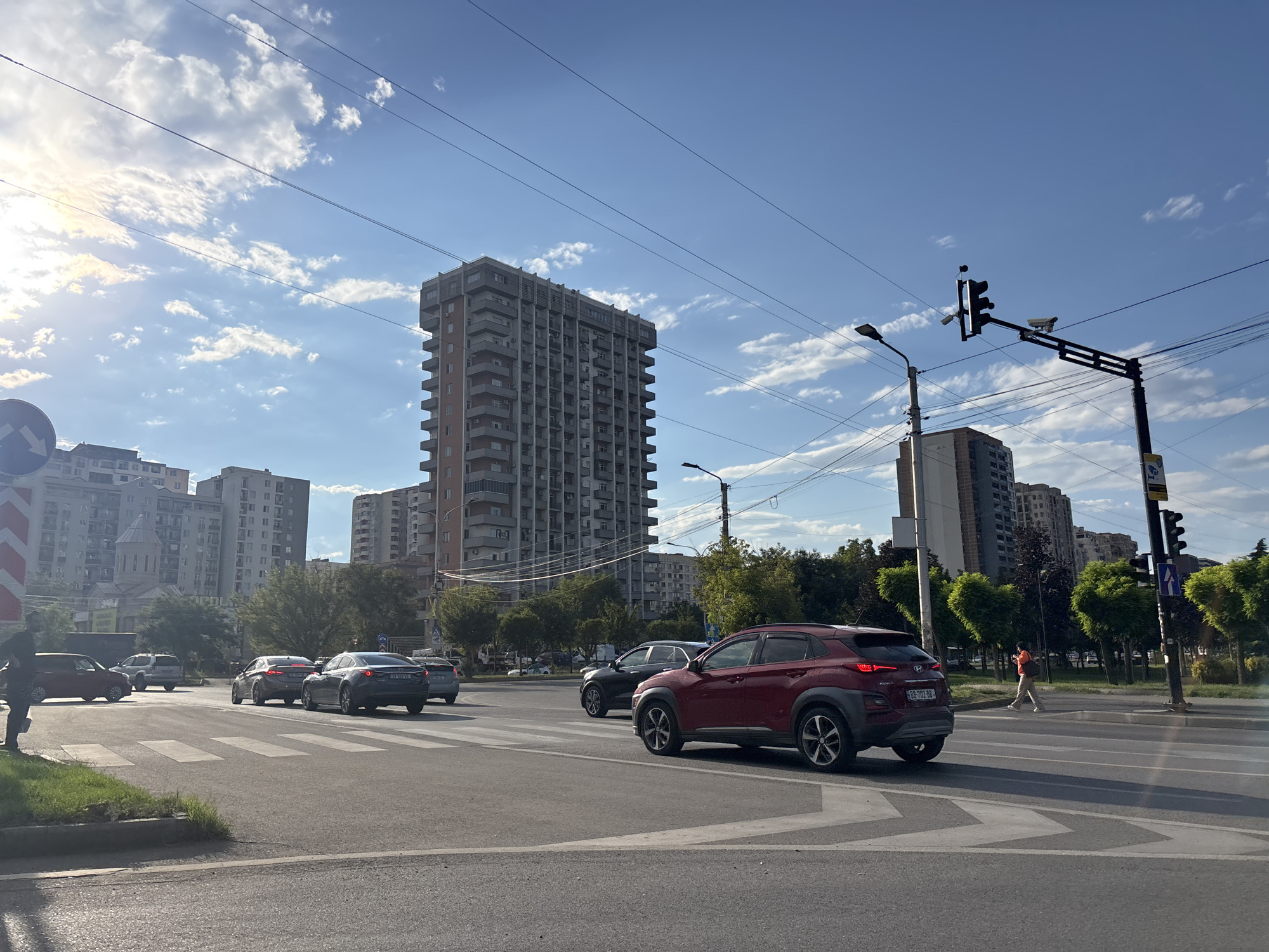 This image captures a street scene in Tbilisi, Georgia, on a sunny day with scattered clouds. The foreground features a pedestrian crossing with several cars. A prominent red SUV is in the right lane, facing away from the camera. Behind it, a dark sedan and a lighter gray sedan are also visible. In the left lane, a dark sedan is visible.
The background is dominated by urban architecture. A tall, Brutalist-style apartment building with numerous balconies stands prominently in the center. To its left and right are other multi-story residential buildings. A traffic light with cameras attached is positioned on a pole to the right of the frame. A street lamp is visible to the left of the traffic light. Trees line the median and the sides of the street, some with distinct heart-shaped canopies. In the far right background, a person in an orange shirt and light-colored pants is walking across a pedestrian crossing. Overhead power lines crisscross the sky. The sun is shining, casting shadows on the road.