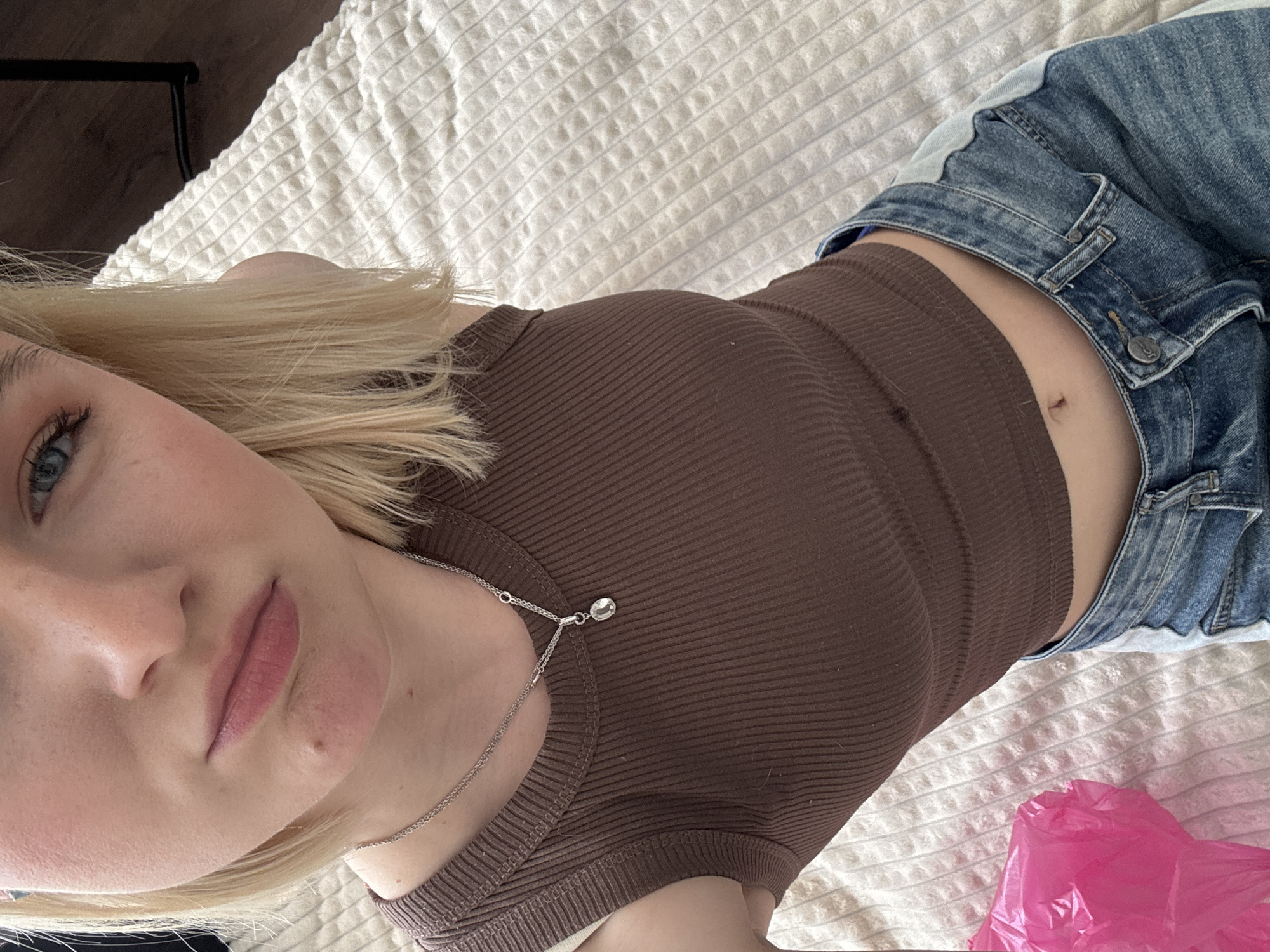 A woman is lying on a bed with a white textured bedspread. She is wearing a brown ribbed tank top and blue denim jeans. A silver necklace with a small pendant is visible around her neck. A pink plastic bag is on the bed near her jeans. The location cannot be determined as Vsevolozhsk, Russia.