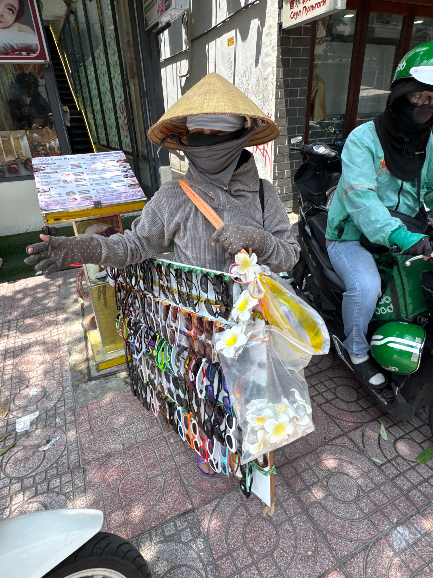 A person wearing a conical hat and a grey hooded sweatshirt is standing on a sidewalk in Nha Trang, Vietnam, selling sunglasses. They are also wearing a grey face covering and grey polka-dotted gloves. In front of them is a display of sunglasses, and to their right is a person on a scooter wearing a turquoise jacket and a green helmet. The sidewalk is paved with patterned tiles, and there is a white scooter in the lower left corner of the image. A menu board for "Lan Spa" is visible behind the person selling sunglasses. Sunlight casts shadows on the ground, indicating a bright day.