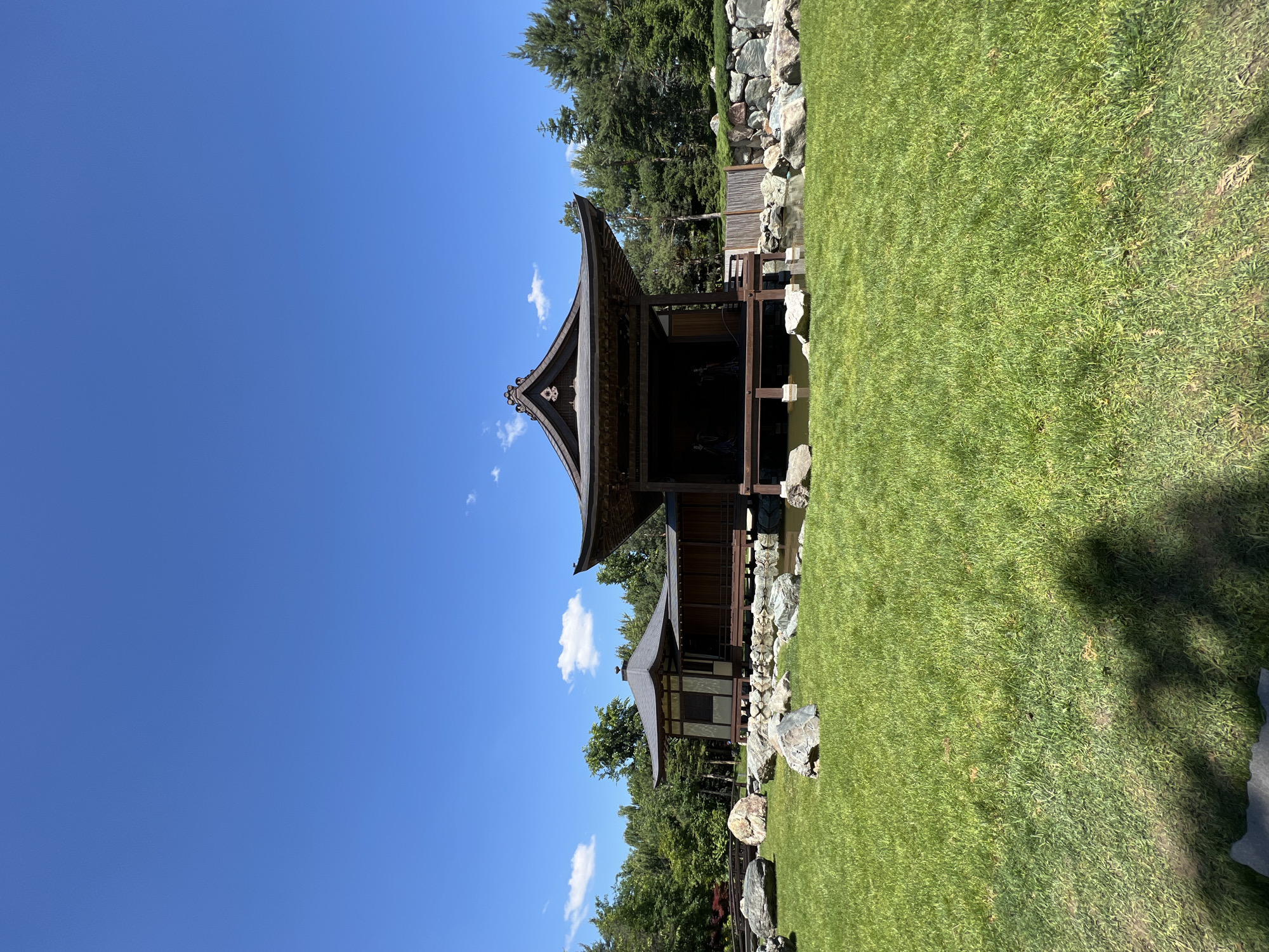 The image displays a serene landscape featuring a traditional Japanese-style pavilion under a clear blue sky. The main pavilion, positioned on the left side of the frame, boasts a distinctive dark, curved roof with an ornate crest at its peak, and appears to be constructed from dark timber, extending partly over a body of water. A smaller, complementary building is visible behind it, partially obscured by trees.
To the right, a wide expanse of well-maintained, vibrant green grass fills the foreground, suggesting a manicured garden or park area. Various rocks are strategically placed along the edges of the grass and near the water, contributing to the naturalistic design. In the background, a line of lush green trees, including some evergreens, rises, framing the scene.
The photograph was taken on a bright, sunny day, indicated by the vivid blue sky, strong lighting, and visible shadows. Only a few scattered white clouds dot the otherwise clear sky, enhancing the peaceful atmosphere. The architectural style and garden elements strongly suggest a Japanese garden setting. Given the context, this tranquil scene is located in Krasnodar, Russia. No specific activities or people are visible, and there is no discernible text in the image.