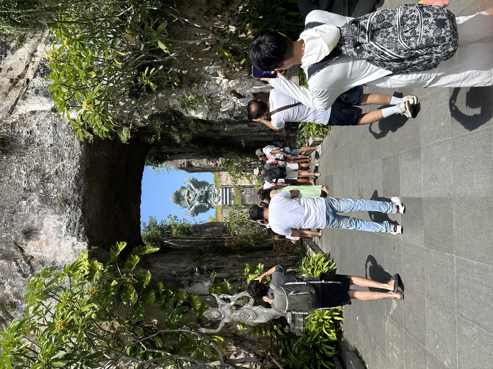 The image depicts a group of tourists visiting a cultural site in Kuta Selatan, Indonesia, likely the Garuda Wisnu Kencana Cultural Park, judging by the large Garuda Wisnu statue visible in the distance. The tourists are standing on a paved pathway, some photographing a stone carving of a deity or mythical figure embedded within a stone wall or archway covered in lush vegetation. The vegetation includes leafy plants and trees, with some bearing small yellow flowers. The tunnel or archway frames the Garuda Wisnu statue, visible against a clear blue sky. The tourists are dressed in a variety of casual clothing, including t-shirts, shorts, and jeans. Several are carrying backpacks or bags. Some are actively taking pictures with their phones or cameras. The scene is well-lit, suggesting it is midday or early afternoon.