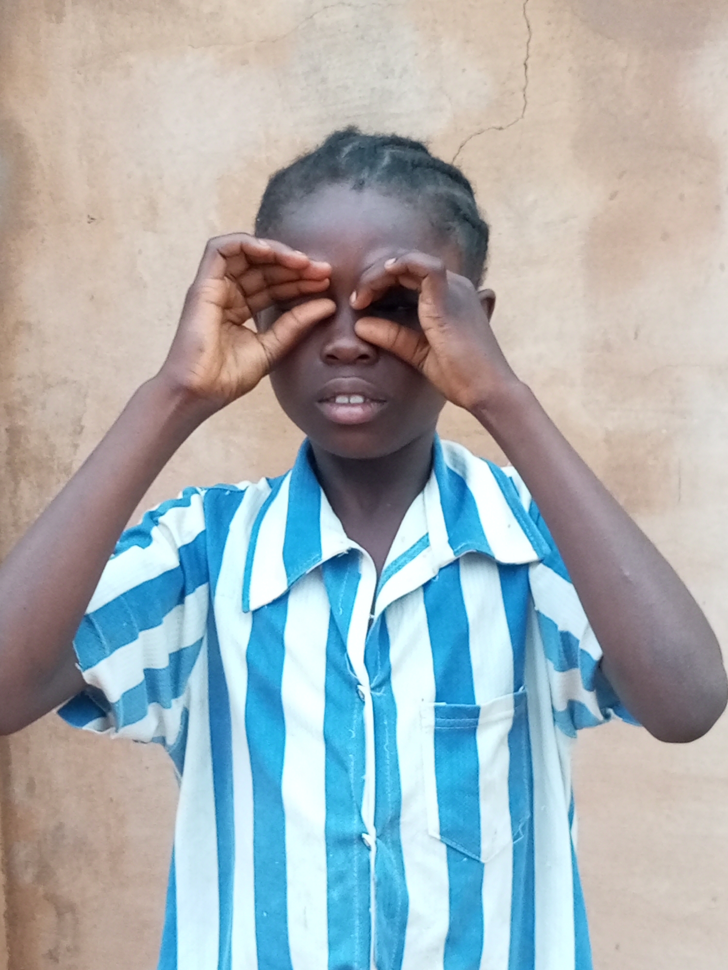 A young person is looking through their hands, which are held up to their eyes like binoculars, against a light brown, textured wall. The person is wearing a blue and white striped collared shirt. Their dark hair is braided. Their mouth is slightly open, revealing their teeth. The background is a plain, unfocused wall with some cracks and imperfections. The overall impression is a candid shot of a child playing or observing something. The image appears to have been taken outdoors in Tempure, Nigeria, indicated by the wall and the appearance of the subject.