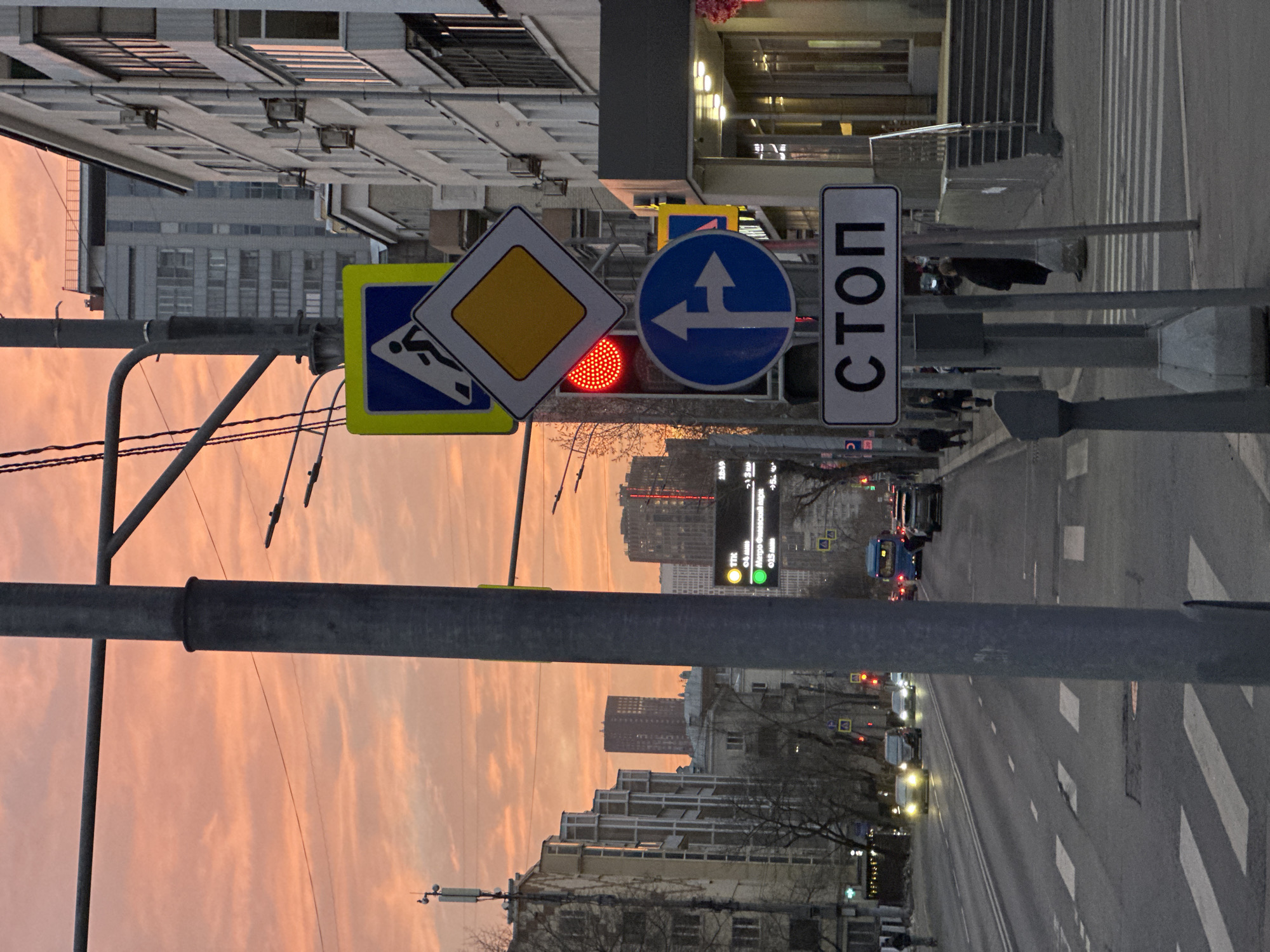 An urban street scene in Moscow, Russia, is depicted at sunset, characterized by a vivid orange and pink sky with scattered clouds. In the foreground, a prominent grey utility pole stands on the right, supporting a vertical array of road signs. These include a blue square pedestrian crossing sign, a yellow diamond representing a priority road, a circular red traffic light, a blue circular sign indicating straight or right turn, and a white rectangular sign displaying "СТОП" (STOP) in black Cyrillic.
A multi-lane asphalt road, marked with white dashed and solid lines, extends into the midground. Several vehicles are present, including cars with illuminated headlights and a blue bus, all positioned away from the viewer. A pedestrian crosswalk with white stripes is visible on the road surface.
Pedestrians are visible on both sidewalks. One individual in dark clothing stands near the utility pole on the right. On the left, an electronic traffic information board displays text in Cyrillic, including "ТТК" (Third Ring Road) and "Метро Филёвский парк" (Filevsky Park Metro Station), along with traffic indicators.
Buildings of varying architectural styles and heights line both sides of the street, with a light-colored multi-story building featuring balconies and external air conditioning units prominent on the right. Bare deciduous trees are interspersed along the sidewalks. In the background, taller, modern high-rise buildings are silhouetted against the sunset, marking the urban skyline.