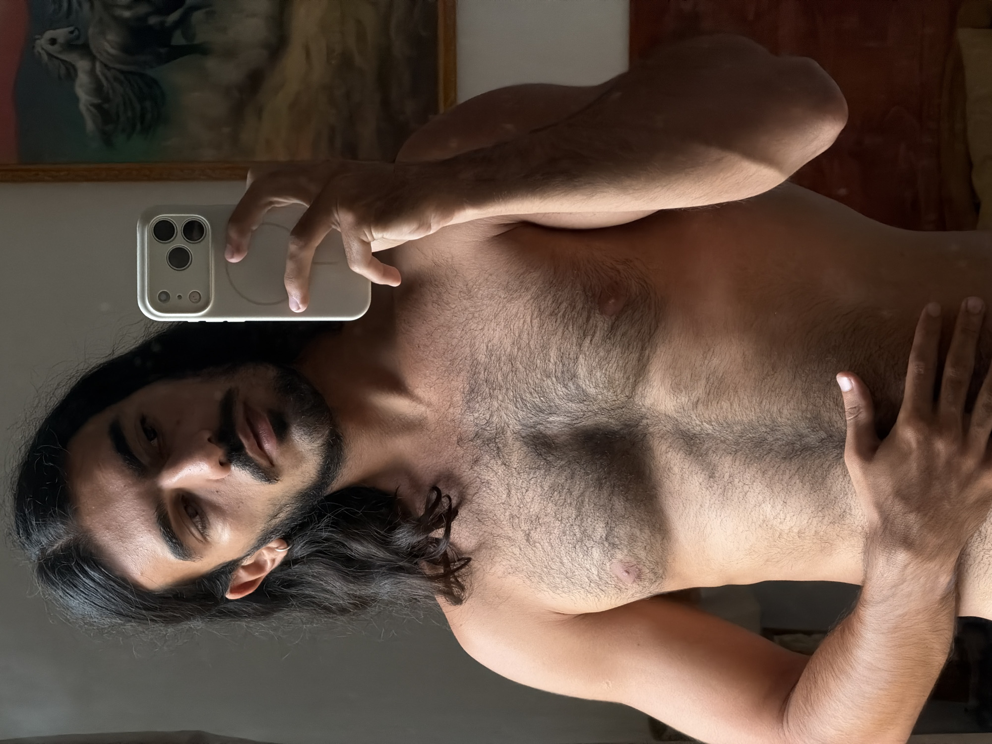 This media file captures a mirror selfie of a man with long dark hair, a beard, and a mustache, taken in North Kuta, Indonesia. The man is shirtless, revealing a visibly hairy chest and abdomen. He holds a light-colored smartphone, likely an iPhone, with a triple-lens camera system and a MagSafe-compatible case, in his right hand. His left hand is placed on his chest.
The man's head is tilted, and he looks directly at his reflection in the mirror with a neutral, calm expression. A small hoop earring is visible in his left ear. The image is oriented with the man's head to the left, indicating it was taken either lying down or rotated.
Reflected in the background behind the man's head is a framed painting, appearing to depict a horse or similar animal in a natural setting. The indoor lighting suggests daytime, casting distinct highlights and shadows across his body, emphasizing his physique and the texture of his hair. There is no visible text in the image.
