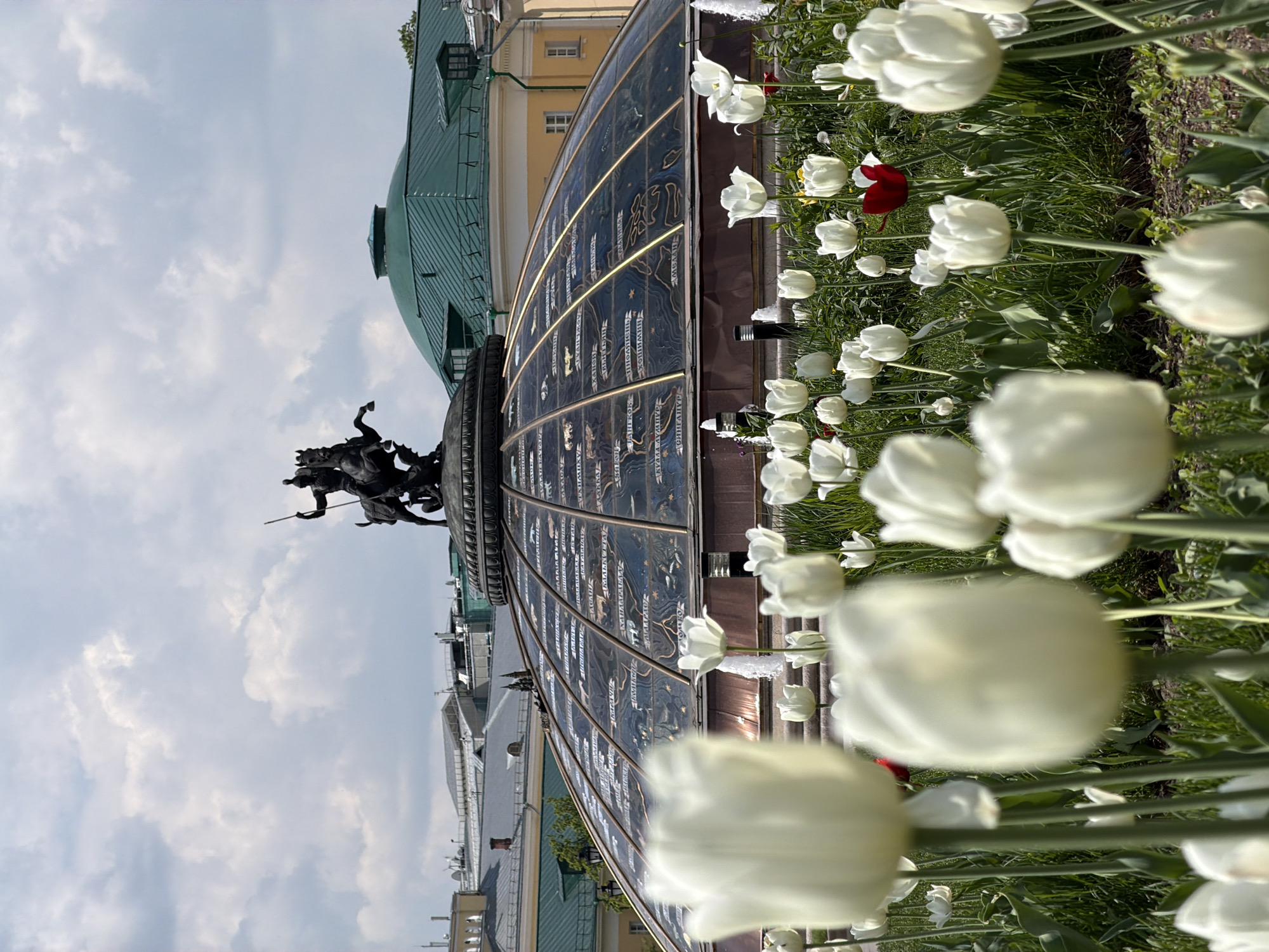 Here is a description of the image:
The image shows the Manegnaya Square fountain complex in Moscow, Russia. The fountain is a large, semicircular structure with a bronze statue of St. George slaying the dragon at the top. The fountain features panels with depictions of the months of the year, along with water jets that create a dynamic display. White tulips are blooming in a flowerbed in the foreground. The buildings of the Moscow Kremlin complex are visible in the background. The sky is partly cloudy.