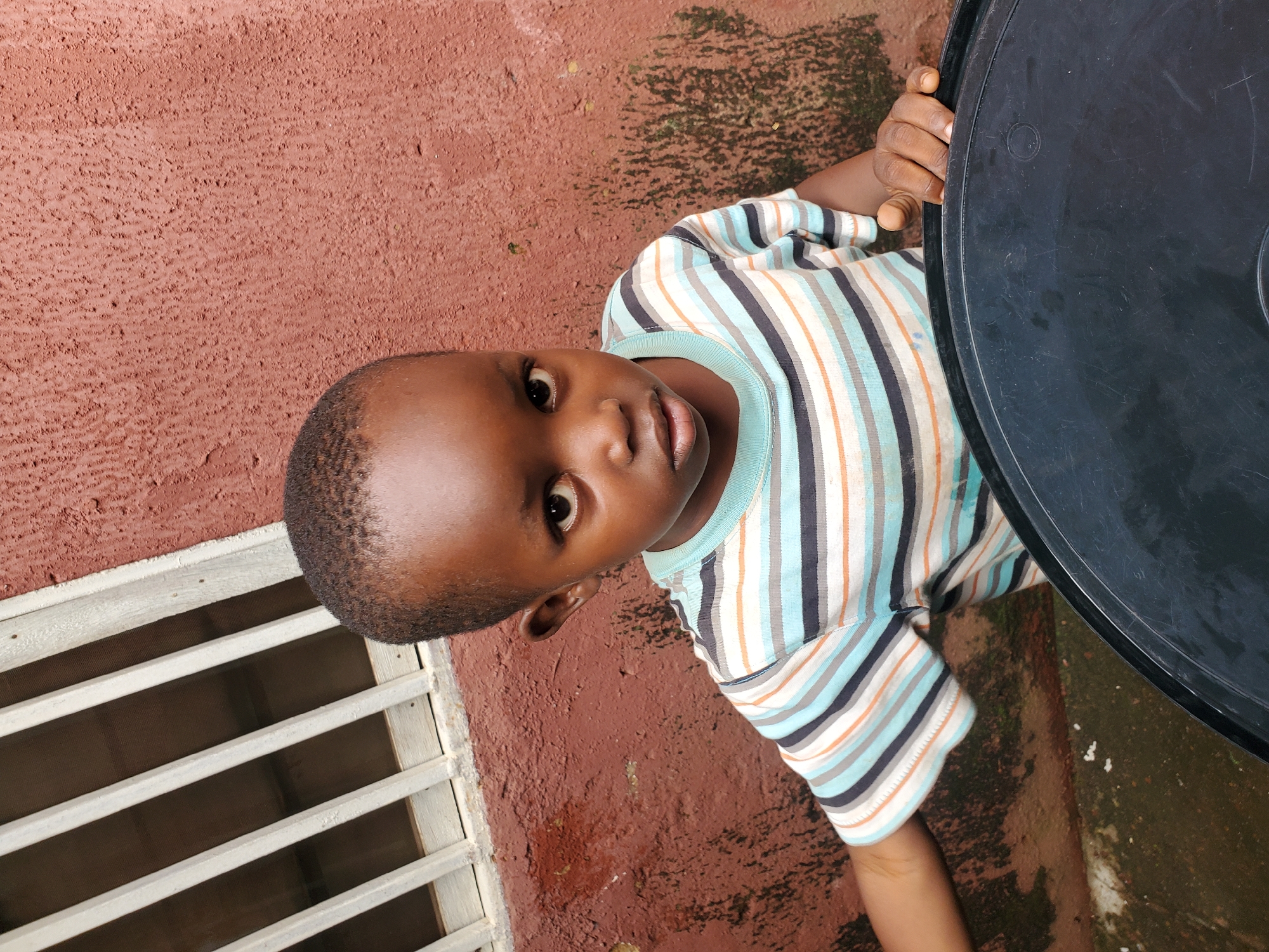 This is an upward-facing medium shot of a young Black child in Jos, Nigeria. The child is looking directly at the camera with a neutral expression. They are wearing a short-sleeved t-shirt with horizontal stripes in shades of blue, white, gray, and orange.
The child is positioned to the left of a large, black, round object, possibly a tabletop, which dominates the right side of the frame. The child's right hand is visible, resting on the edge of this object.
The background consists of a textured, reddish-brown wall, appearing to be concrete or plaster. To the left of the child, there is a window with horizontal white bars and a dark mesh screen behind them. The ground is a mix of reddish earth and some darker, possibly wet or mossy patches. The lighting suggests it is daytime, with no distinct shadows indicating a particular time. The overall scene appears to be outdoors in a simple, possibly residential, setting. There is no visible text in the image.