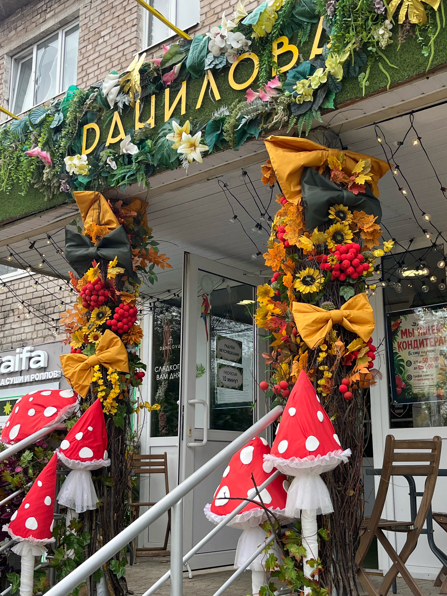 The image shows the storefront of a cafe, likely named "DANAILOVA" judging by the Cyrillic lettering on the facade. The entrance is decorated with an autumnal theme, featuring artificial foliage, sunflowers, red berries, and large yellow and dark green bows. Red and white polka-dotted mushroom figures are arranged on the steps leading up to the entrance, some with white tulle skirts. String lights are hung above the entrance, and a sign in Russian is visible on the right side of the door. A portion of a sign for a sushi and roll shop, "Raifa," is visible to the left. The building appears to be made of brick, and wooden chairs and tables are visible outside the cafe. The overall impression is one of a festive and whimsical autumnal display.