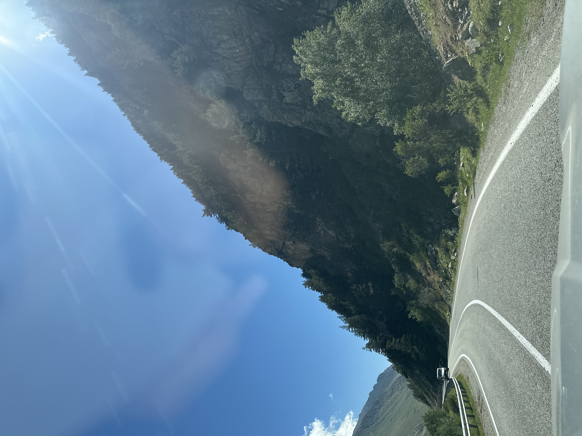 The image depicts a winding paved road through a mountainous region, likely in or near Upper Baksan, Russia. The road curves to the left, with a metal guardrail on the left side. A white van is visible driving away on the road in the distance. The right side of the road is a steep, rocky mountainside covered with dark green coniferous trees and some deciduous trees. The sun is shining brightly from the upper right corner of the frame, creating lens flares and casting light and shadows on the mountain. The sky is clear and blue. The foreground shows the top of a vehicle's dashboard. The overall scene suggests a scenic drive in a rugged, natural environment.