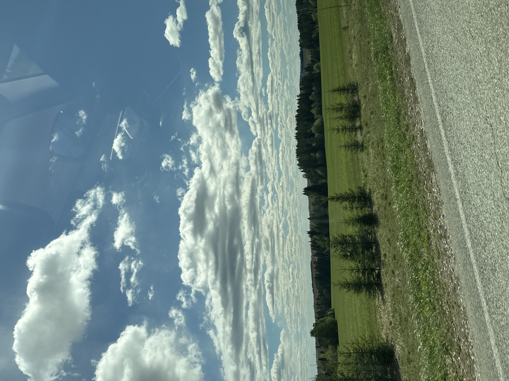 The image shows a view from a vehicle, likely a car, while traveling on a road. The sky is visible through the front window, exhibiting a blue expanse populated with cumulus clouds. To the right of the frame, a landscape features a green field bordered by a line of trees, adjacent to a section of asphalt road. The interior of the vehicle is partially reflected in the glass. The location is presumably near Ledukhi, Russia, based on the prompt, though this cannot be definitively determined from the image alone.