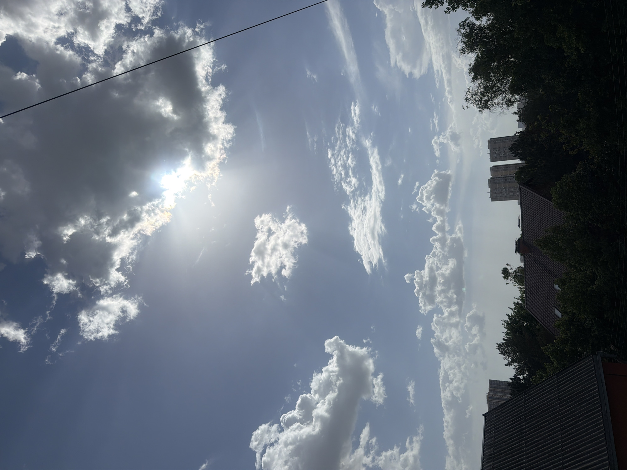 The image depicts a bright, daytime sky with scattered clouds. The sun is visible behind one of the larger cloud formations, casting a glare. A power line is visible in the upper left corner. In the lower right, rooftops and trees are visible, with two high-rise buildings appearing in the distance. The location is identified as Rostov-on-Don, Russia.