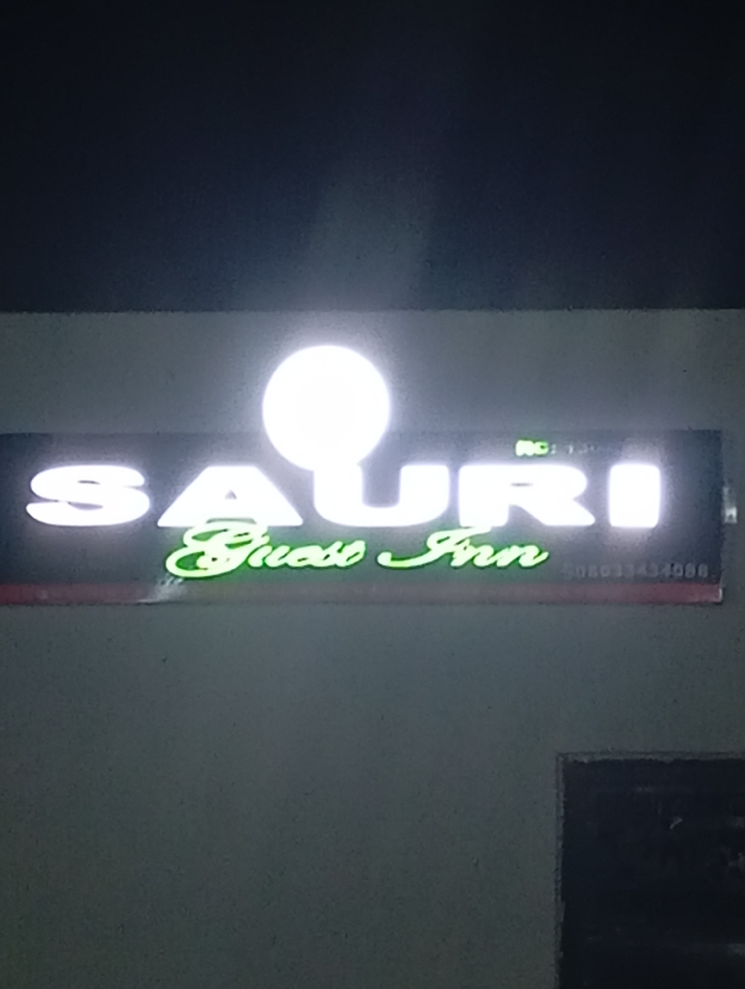 A horizontal, rectangular illuminated sign is affixed to a dark gray, textured wall under low-light conditions, likely at night. The sign displays two lines of text. The upper line, "SAURI," is rendered in large, capital, glowing white letters. A bright, circular light source or glare partially obscures the middle portion of "SAURI," specifically over the 'U' and parts of the adjacent letters. Above the letters "URI," faint green text reads "NO: 130." The lower line of text, "Guest Inn," is presented in glowing green cursive script. To the right of "Guest Inn," a sequence of numbers, "08033434088," is faintly visible in small text on the sign's dark background. The wall surrounding the sign appears uniformly dark and textured, providing a stark contrast to the illuminated sign. The location is Unknown, Unknown.