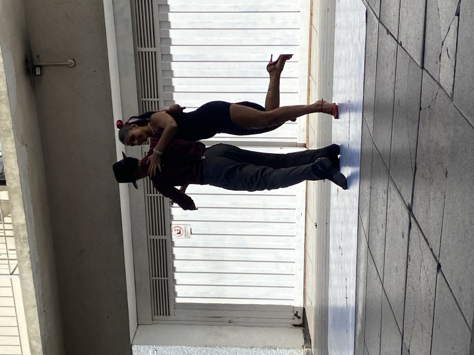 Two individuals, a man and a woman, are engaged in a dance outdoors in a public space in Buenos Aires, Argentina. The male subject wears a dark fedora, a long-sleeved maroon shirt, grey trousers with suspenders, and dark shoes. He is positioned with his back partially to the viewer, his left arm around the woman's waist. The female subject wears a dark, short dress, pink high-heeled shoes, and has her hair tied back with a red accessory; she has multiple bracelets on her right wrist and is smiling, looking towards her partner with her right leg raised backward. Her right arm rests on the man's shoulder. They are dancing on a light-colored square mat placed over a dark, cracked concrete tiled sidewalk. The immediate background features a white corrugated garage door with horizontal ventilation slats and a "No Parking" (E) sign affixed to it. Above the garage door is a concrete overhang with a metal railing. A light grey stucco wall with the number "144" is visible on the far left. The scene is illuminated by natural light.