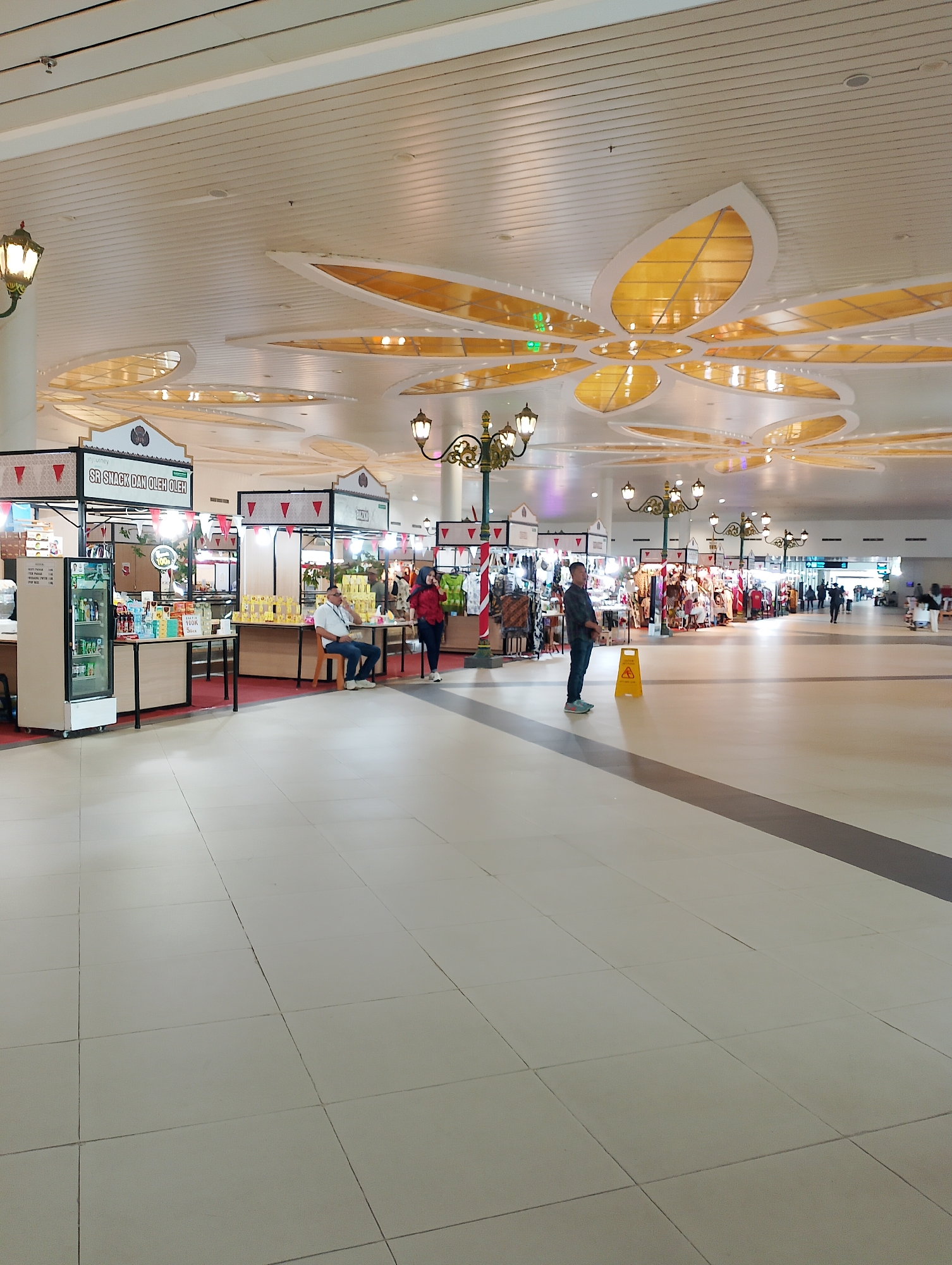 The image depicts a spacious, indoor marketplace with an arched ceiling adorned with floral-shaped lighting fixtures. The floor is tiled in a light color, and the stalls are set up along the sides of the walkway.
In the foreground, a man in a plaid shirt and jeans stands facing right, next to a yellow wet floor sign. To his left, a refrigerated display case containing bottles of drinks and various food items is visible.
Further into the marketplace, several stalls are set up. One stall has a sign that reads "SR SNACK DAN OLEH OLEH". A man in a white shirt and dark pants sits on a chair in front of one of the stalls, looking down. To his right, a woman in a red shirt and dark pants walks away from the camera.
Other stalls display clothing and various merchandise. In the background, more people can be seen walking through the marketplace, and a long corridor stretches into the distance, with more stalls and lighting fixtures lining the walls. The overall atmosphere suggests a busy commercial area. The location is identified as Palihan, Indonesia.