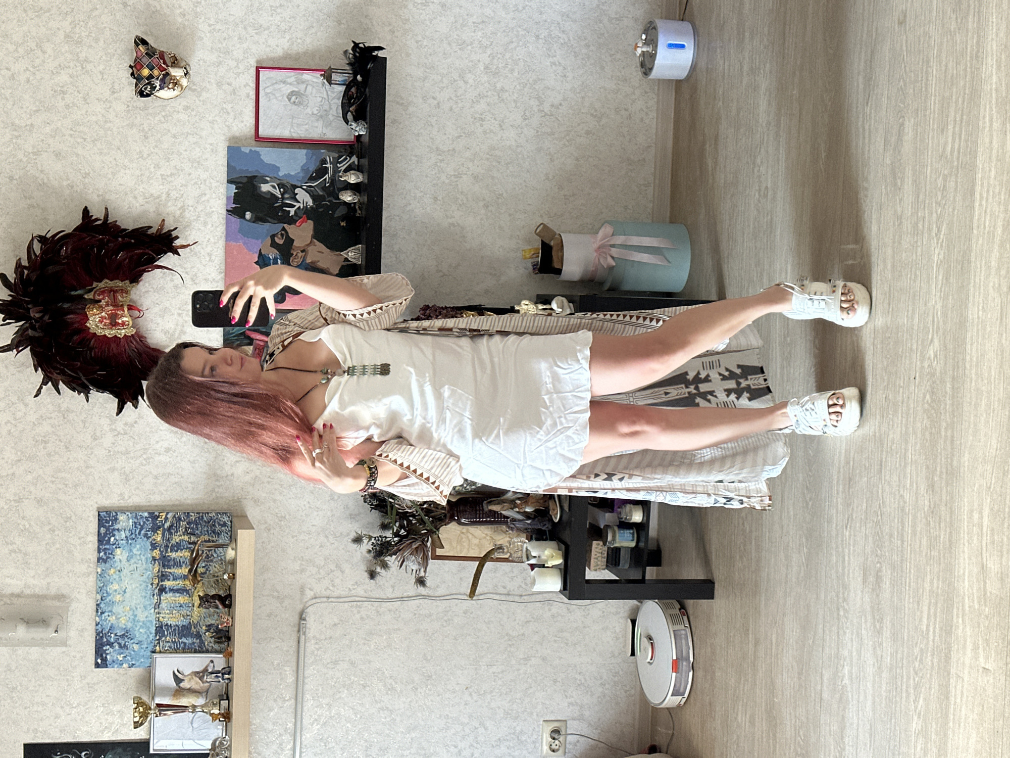 A young woman with long, reddish-brown hair stands indoors, taking a mirror selfie with a black smartphone. She is wearing a light-colored sleeveless dress or top, a long patterned cardigan, and white chunky sandals or sneakers. Her accessories include rings, a bracelet, and a necklace. Her expression, visible on the phone screen, appears neutral or slightly smiling.
The background reveals a decorated living space. On the left, a wall features a colorful Venetian-style mask and a large red and gold feathered decoration. Below these, a wooden shelf displays a gold trophy, small figurines, and a framed artwork of a deer. Another painting, possibly depicting Batman, is visible. The walls are light-colored with a subtle texture, and the floor is light wood laminate.
To the right, a light-blue gift box with a pink ribbon is present, along with a black shelving unit holding various decorative items, including small bottles, candles, and artificial plants. A white robotic vacuum cleaner is on the floor near a power outlet. Two small devices, one light-blue and cylindrical, and another white, are mounted on the wall. The setting suggests a casual, contemporary home environment during daytime. No discernible text is visible in the image. The location context for this scene is Vladimir, Russia.