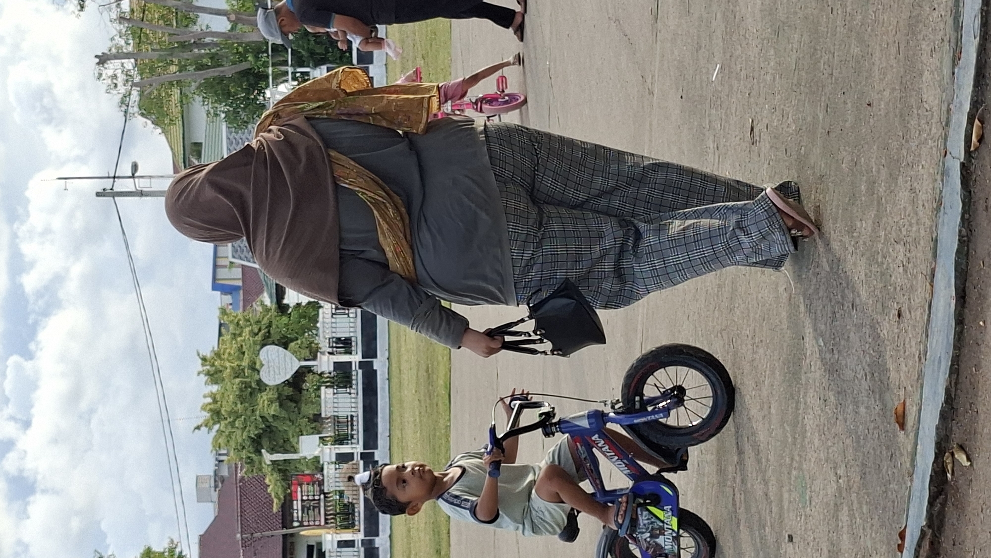 In Lhokseumawe, Indonesia, a young boy rides a blue and black "MONTANA" bicycle on a paved surface. He wears a light grey t-shirt and shorts, and appears to be looking to his left. Walking to the right of him, and slightly behind, is an adult wearing a brown hijab and a grey long-sleeved shirt. She also wears plaid pants and brown flip-flops, and carries a black handbag in her left hand. A yellow scarf is draped over her right shoulder. In the background, a child on a pink bicycle is visible, as well as other people and buildings. The sky is cloudy.