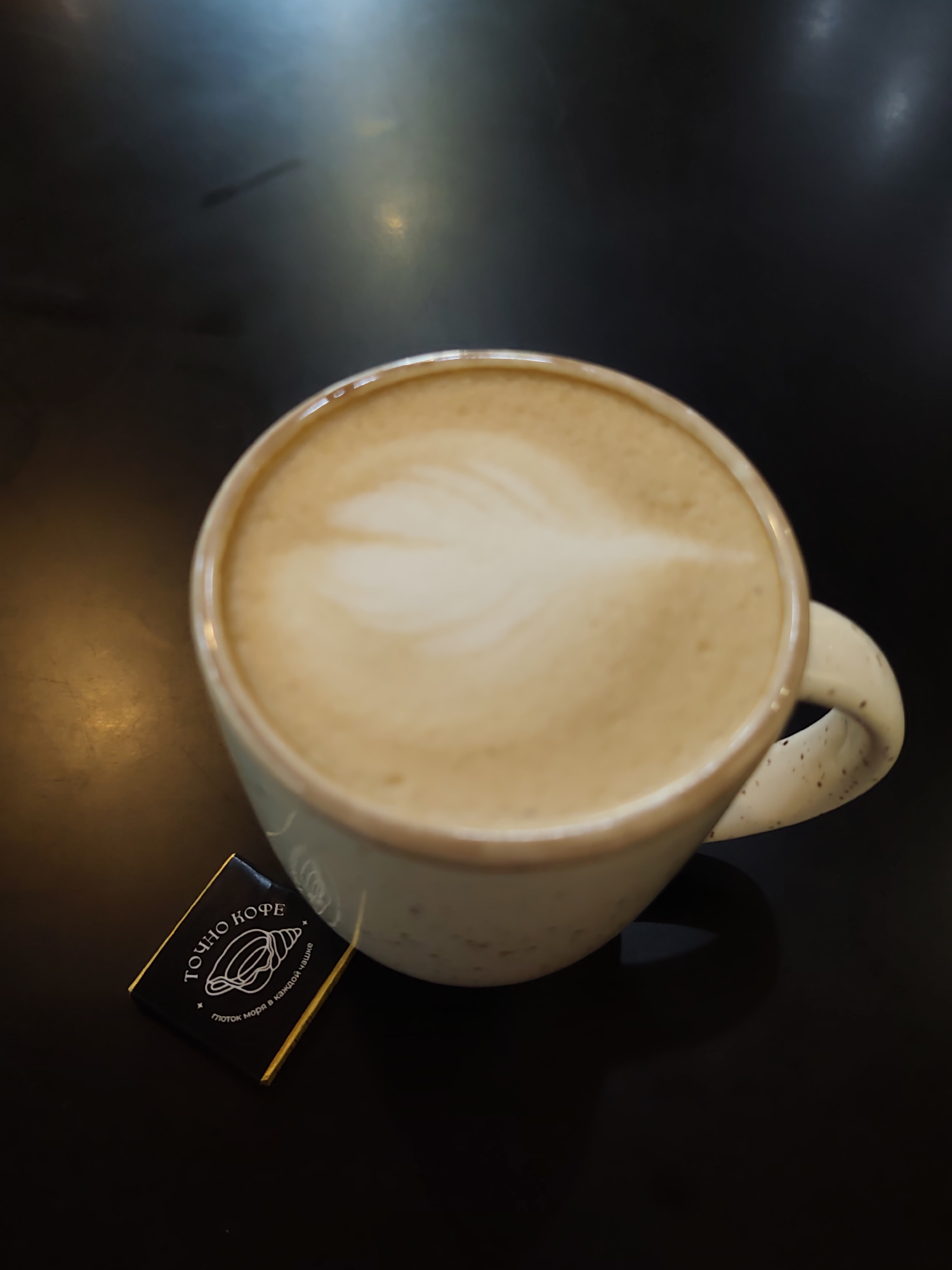 An overhead shot shows a cup of coffee with latte art on a dark, reflective surface, likely a table. A small, dark chocolate square with white text and a seashell illustration is positioned to the left of the cup. The chocolate wrapper reads "ТОЧНО КОФЕ" (Exactly Coffee) and "Глоток моря в каждой чашке" (A sip of the sea in every cup). The coffee appears to be a cappuccino or latte, with a foamy, milky-brown surface and a delicate leaf-like design on top. The cup is light-colored with small brown speckles and a curved handle. The background is dark with subtle reflections of light, indicating an indoor setting. There are no people visible in the image. The overall impression is one of a cozy, possibly cafe setting, in Sochi, Russia.