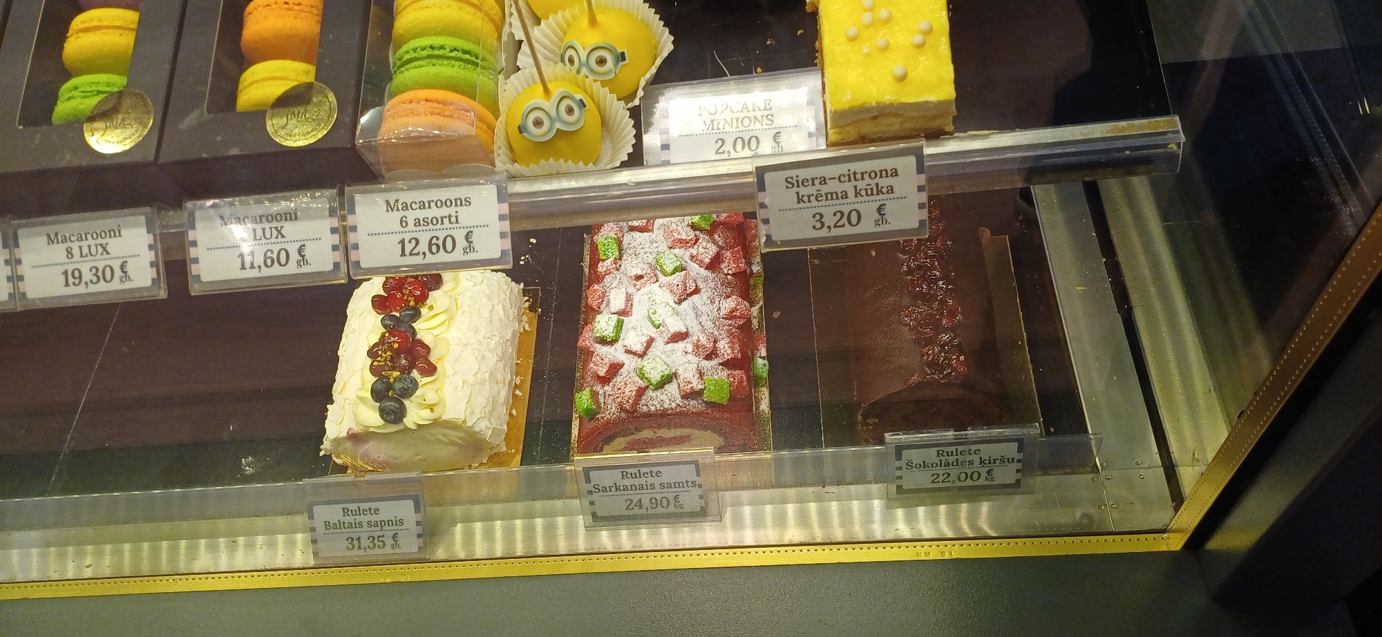 This is a close-up, eye-level, slightly angled, interior shot of a bakery display case filled with various desserts and pastries. The lighting is bright, illuminating the products and creating reflections on the glass.
In the foreground, on the left, is a white cake roll, "Rulete Baltas sapnis", priced at 31.35€. To its right, another cake roll, "Rulete Sarkanais samts", is visible, priced at 24.90€. Below that, a dark chocolate cake roll, "Rulete Šokolādes kiršu", is priced at 22.00€.
Further back, in the center, are macarons in boxes, with labels indicating "Macarooni 8 LUX 19,30€", "Macarooni 8 LUX 11,60€", and "Macaroons 6 asorti 12,60€".
On the right side of the display, there are yellow cake pops decorated to resemble Minions, with a price tag of "PUPE 2,00€". Next to them are colorful macarons in a plastic box. Another cake slice, yellow with white icing dots, is visible at the bottom right, with a label reading "Siera-citrona krēma kūka 3,20€".
The overall environment appears to be a professional food display, likely in a store or cafe, with a focus on visually appealing, freshly made desserts. There are no people or actions explicitly captured in the image. The background is dark and indistinct, suggesting the focus is solely on the merchandise.