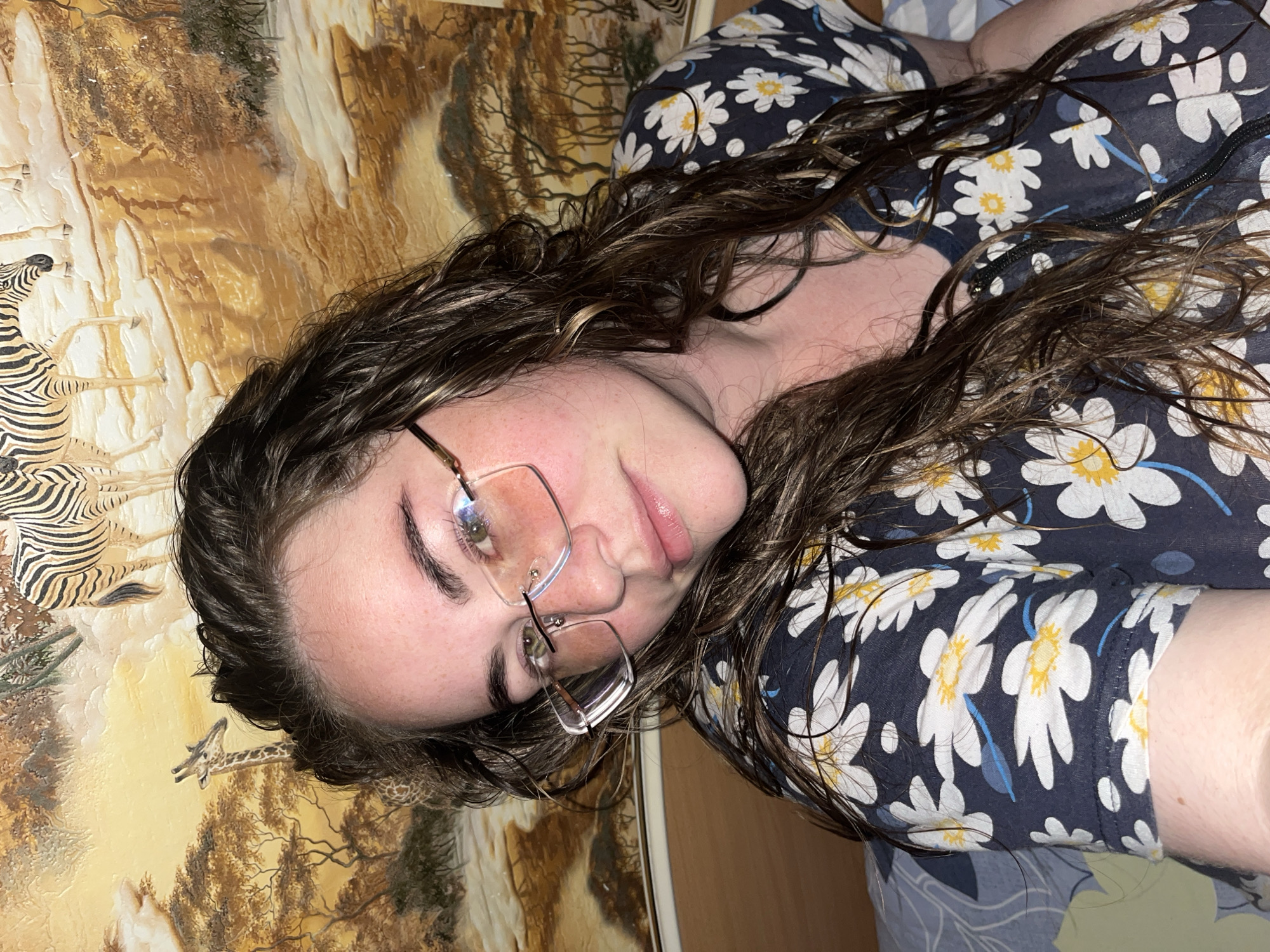 A close-up photograph depicts a female subject indoors in Сосново, Russia. The subject has long, dark brown, wavy hair that appears somewhat wet or styled. She has fair skin with visible freckles and wears rectangular eyeglasses with thin metal frames, exhibiting a slight pink reflection in the lenses. Her eyes, which appear greenish-brown, are directed towards the viewer. She is wearing a dark blue top adorned with white and yellow daisy patterns and light blue floral accents. The top has a visible neckline with what appears to be a zipper or seam. The background features a wall or headboard decorated with an African safari-themed mural, rendered in warm yellow and brown tones. The mural depicts zebras, a giraffe, and savanna trees. A light brown wooden structure, possibly a headboard, is visible behind the subject's left shoulder, and a patterned blue and white fabric, likely bedding, is visible in the lower left corner.