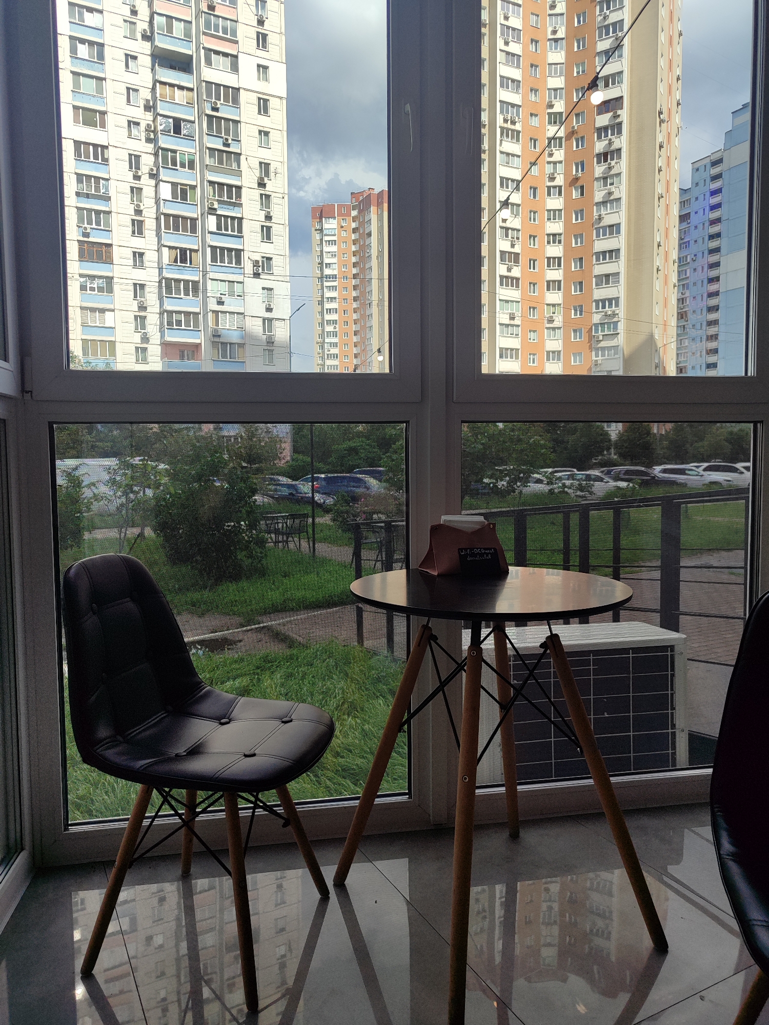 In a brightly lit room with a reflective tiled floor, a black leather upholstered chair with wooden legs is positioned to the left. In front of the chair, a dark, round table with three wooden legs stands. A small brown holder with napkins and a chalk sign rests on the table. Beyond the table and chair, a large window reveals an outdoor scene. Tall, multi-story residential buildings with numerous windows and air conditioning units are visible in the distance, under a cloudy sky. The foreground of the outdoor view shows a grassy area with trees and parked cars. An air conditioning unit is visible outside the window, below the table.