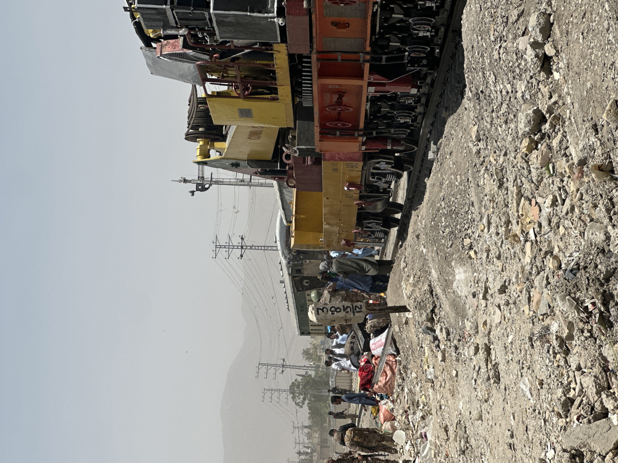 In Quetta City Tehsil, Pakistan, an out-of-focus foreground depicts a rocky, trash-strewn area. Along railway tracks, a train comprised of multiple yellow and red carriages is stopped, with a group of civilians alongside. Some people are sitting, while others are standing near their belongings, which appear to be wrapped in colorful fabrics. A soldier in military fatigues is among the crowd. Power lines are visible in the background, stretching across the landscape towards mountains shrouded in haze. The sky is overcast, contributing to an overall muted tone in the scene.