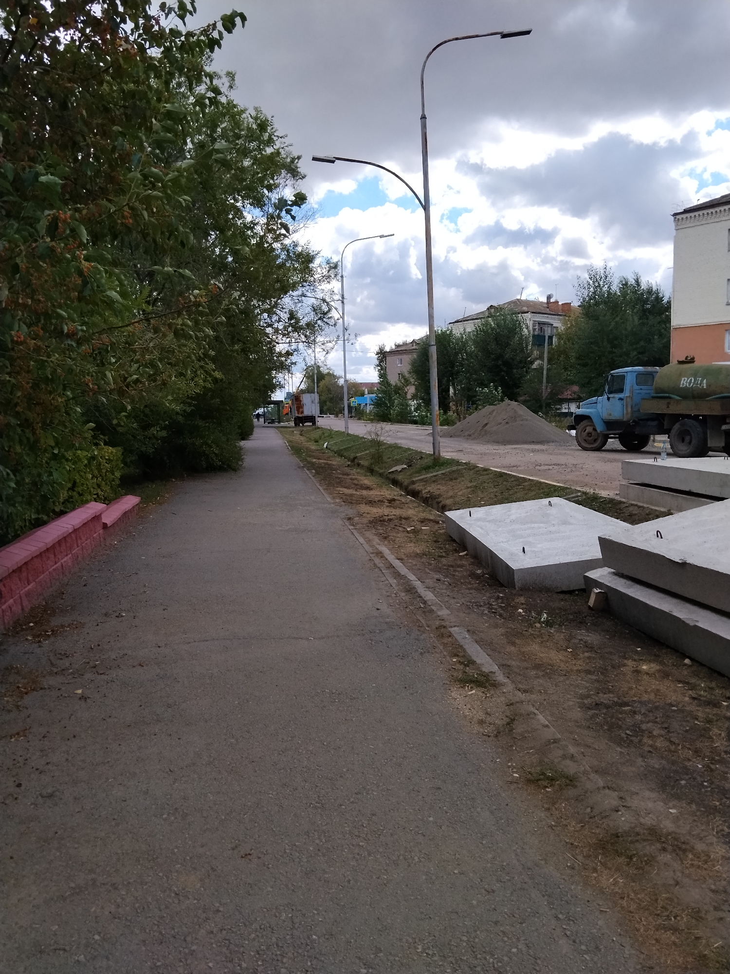 The image depicts a street scene in Kokshetau, Kazakhstan, with construction materials and vehicles present. On the left, a paved sidewalk runs alongside a dense row of green trees. The sidewalk is bordered by a low, pink-painted concrete wall. Across the street, a pile of sand is visible, with a blue truck parked nearby. The truck has a large tank on its back, labeled "ВОДА" (water). Several large, rectangular concrete slabs are stacked on the right side of the road, indicating ongoing construction or repair work. Streetlights are present along the sidewalk. The sky is overcast with scattered clouds and patches of blue sky. The overall impression is of a street undergoing infrastructure maintenance.