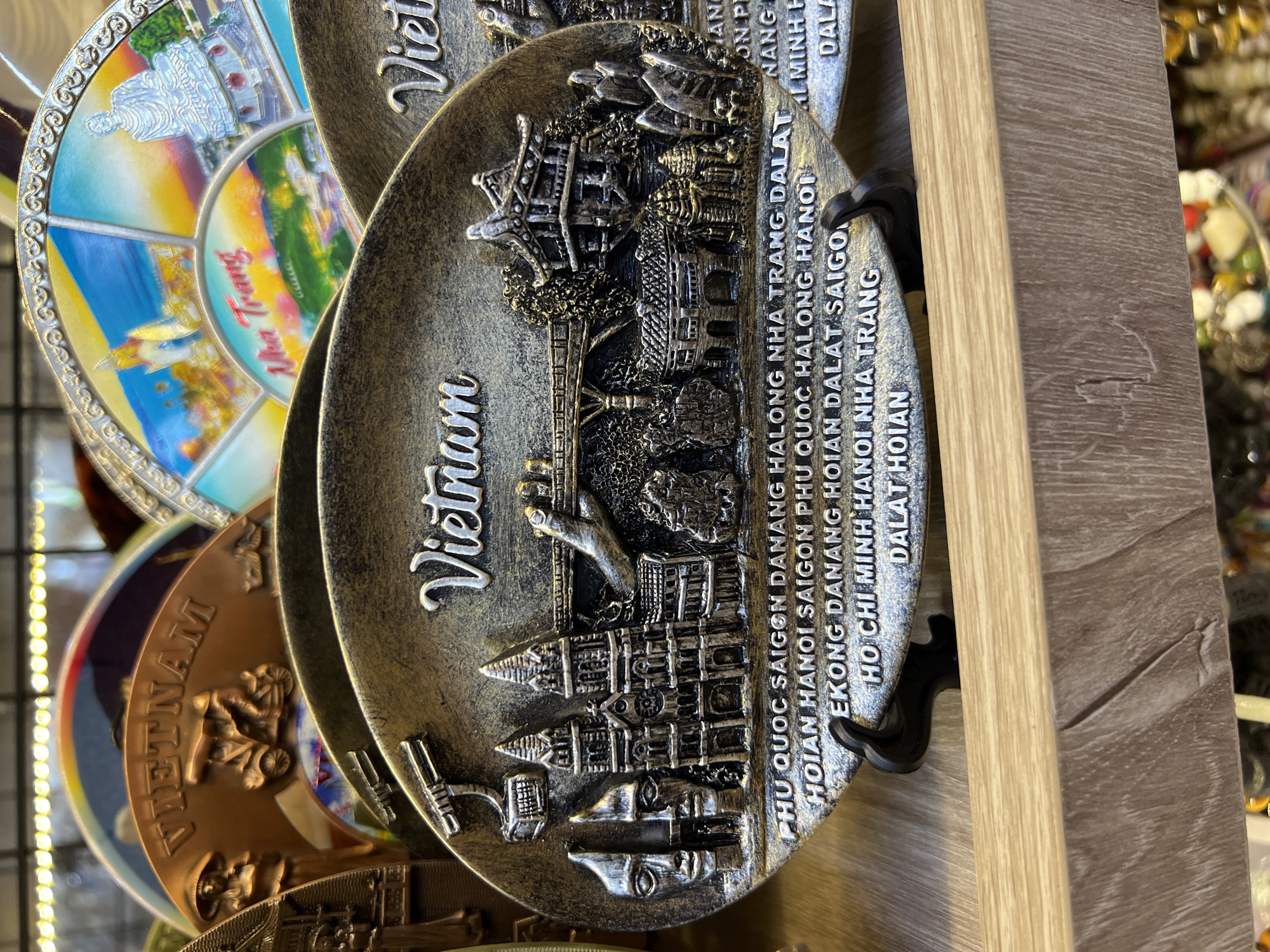 The image shows a close-up of several souvenir plates, likely for sale in a tourist shop. The plates are embossed with images of Vietnamese landmarks and text. The central plate, which is the most in focus, has "Vietnam" written across the top and depicts a cityscape with several buildings, a bridge, and a statue of a hand. Below the cityscape, there is text listing various Vietnamese cities. The plate is metallic and appears to be a dark bronze or antique gold color. It is displayed on a black stand in front of other similar plates. The background is blurred but shows more souvenir items, suggesting an indoor retail environment. One of the other plates visible is a fan-shaped souvenir with a depiction of a pagoda and a large Buddha statue, with "Nha Trang" written in the center. This suggests that the souvenirs are related to popular tourist destinations in Vietnam, including Nha Trang. The overall scene is a retail display of tourist memorabilia.