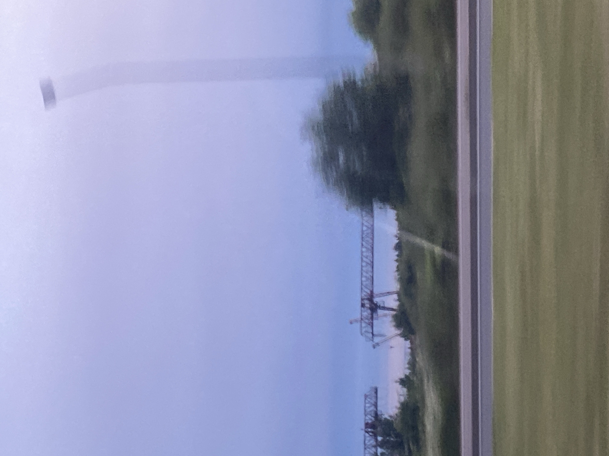 This media file captures an outdoor scene with a hazy, muted sky. The left side of the frame is dominated by a soft, pale blue atmosphere, suggesting a cloudy or foggy day, or perhaps early morning or late evening. A faint, elongated, light-colored object, possibly a distant tower or structure, is visible in the upper left quadrant, blurred by motion or atmospheric conditions.
To the right of the hazy sky, a distinct tree line with dark green foliage forms a dense border. Beyond this, a patch of bright green, striped ground, likely a field or mowed area, runs along the far right edge of the image. A vertical element, possibly a pole or thin structure, stands starkly against the sky in the far left of the frame.
In the middle ground, a complex, dark-colored industrial structure, resembling a gantry crane or a section of a bridge under construction, is visible. This structure is segmented and appears to be made of metal beams. Further into the background, another similar, but smaller, dark structure can be discerned.
The overall impression is one of a somewhat industrial or construction landscape viewed through a veil of atmospheric conditions, potentially from a moving vehicle given the blurriness. The lighting is diffused, indicating neither bright sunlight nor deep darkness. There is no visible text.