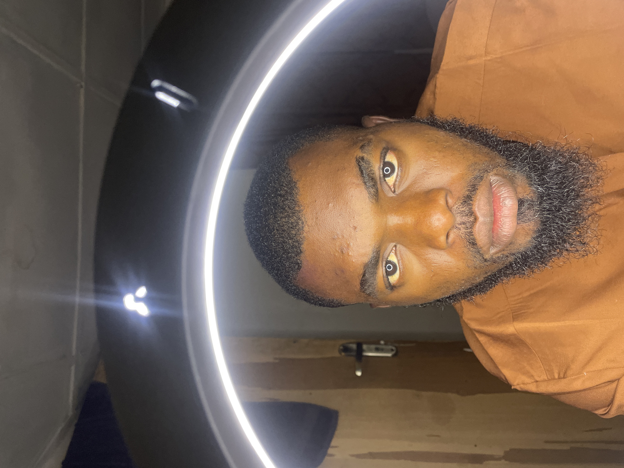 The image displays a close-up, rotated portrait of a dark-skinned man with a neat haircut and a well-groomed, dark beard. He is wearing a plain, long-sleeved, caramel-colored shirt. His face is brightly lit by a circular ring light, which is partially visible on the left side of the frame and creates distinctive bright circular reflections in the pupils of his widely open, yellowish eyes.
The man gazes directly forward with a neutral to intense expression. His skin shows some texture and appears slightly shiny, with minor blemishes on his forehead and cheek. His lips are full and have a reddish hue.
The background is indistinct, suggesting an indoor setting. On the left, there is a light-colored wall or surface, while on the right, a wooden surface, possibly part of a door or furniture, with a small metallic fixture is visible. The dominant light source is the artificial ring light, indicating a studio setup or well-lit indoor environment. No specific time of day or weather can be discerned from the scene. There is no visible text. The location context provided for this image is Jos, Nigeria.