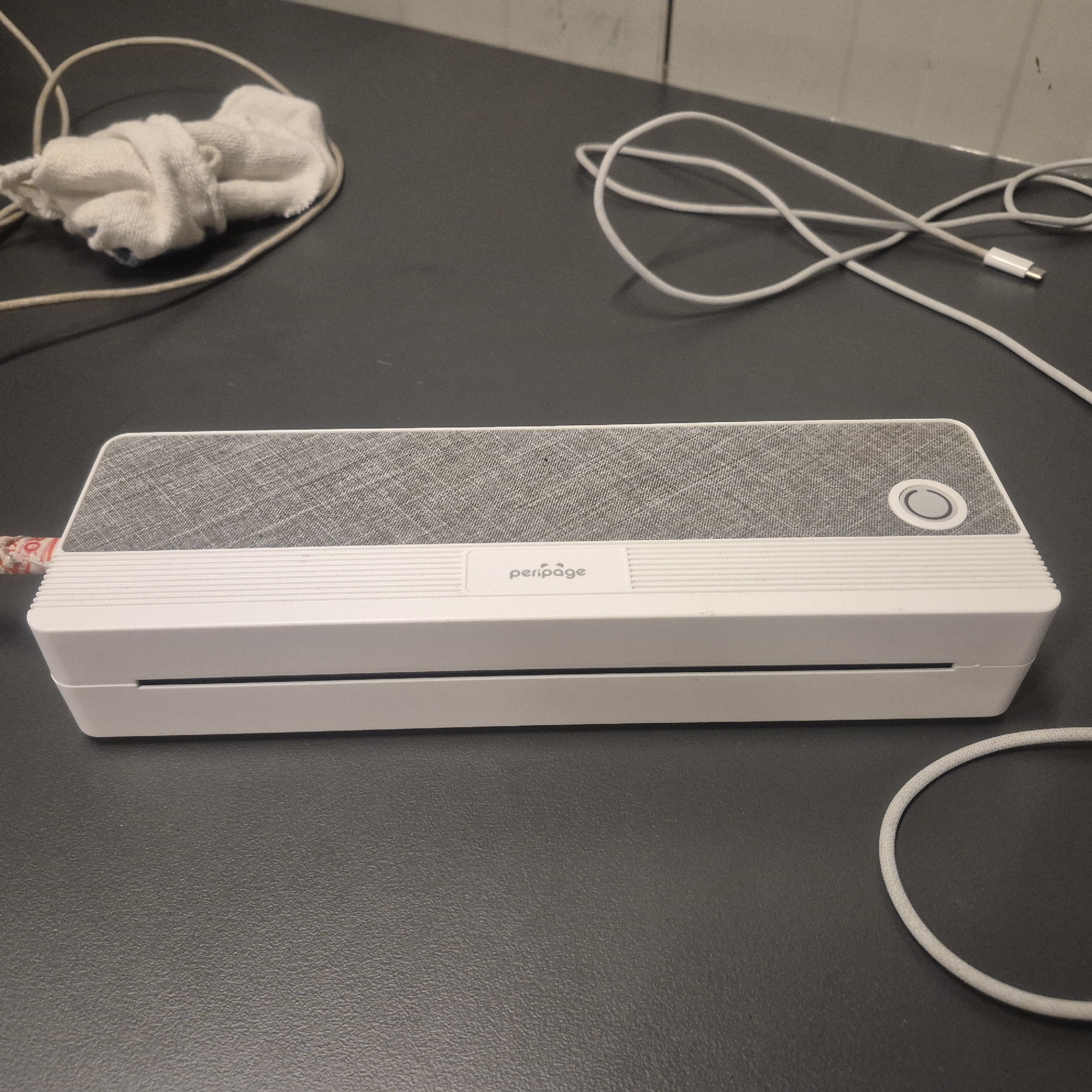 A white and grey Peripage printer is situated on a dark grey textured surface, possibly a desk. The printer is elongated and has a patterned grey top with a circular button on the right side. Below the grey section, there are horizontal white ridges and a slot at the bottom. A white USB-C cable is connected to the left side of the printer and extends across the surface, looping around and then leading out of frame to the right. A white crumpled cloth or sock is also visible near the left side of the printer, with a light brown string wrapped around it. The background of the image shows a light-colored wall with some faint marks. The scene is indoors, and there are no people present.