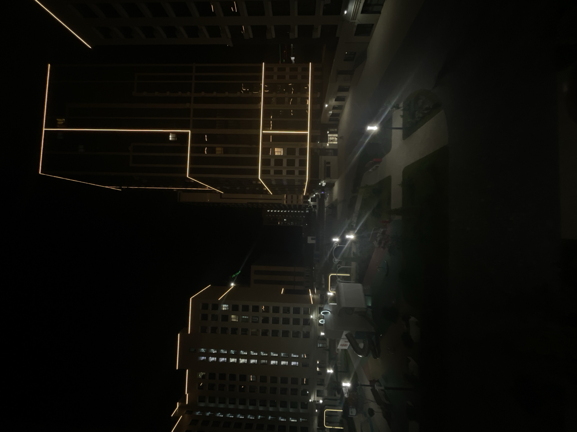 The image captures a nighttime view of a modern residential complex in the city of Stavropol, Russia. The scene is dominated by tall, multi-story buildings with illuminated outlines in warm yellow light, accentuating their geometric designs. The buildings feature numerous windows, some of which are lit from within, indicating occupancy.
In the foreground and mid-ground, a courtyard or public space is visible, equipped with streetlights casting pools of light on paved pathways and grassy areas. Playground equipment, including a slide and other structures, is present in the courtyard, suggesting a family-friendly environment. The overall atmosphere is one of urban development and nighttime illumination. There are no people visible in the image. The environment appears to be clean and well-maintained.