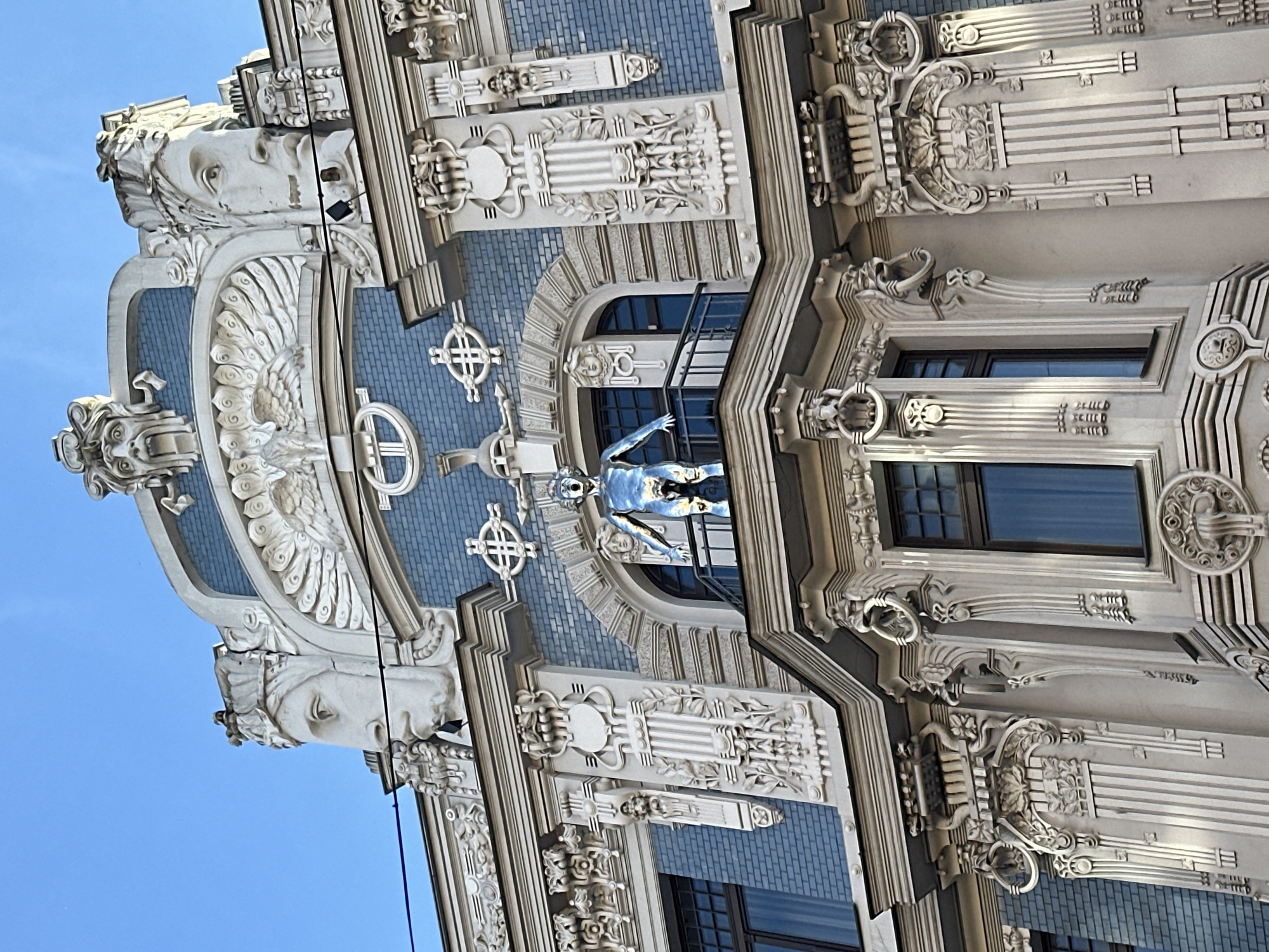 An upward-angled view captures the ornate facade of an Art Nouveau building in Riga, Latvia, under a clear blue sky. The architecture is highly decorative, featuring numerous sculptures and intricate relief work.
Prominently featured are large, intricately carved white busts of women, positioned at the upper corners of the depicted section of the building. Their faces convey a serene or classical expression, framed by flowing hair. Below them, a large, stylized bird-like figure with prominent, fanned wings, possibly an eagle or owl, is central to the design. This creature is set against a light blue, brick-patterned background, surrounded by other geometric and organic decorative elements.
Further down, within a recessed balcony area, stands a striking, silver-colored statue of a human figure, possibly male, with arms outstretched. This figure has a smooth, metallic sheen, contrasting with the stone of the building. Surrounding it are additional carved details, including smaller busts and stylized patterns.
The facade is rich with decorative motifs, incorporating floral elements, geometric shapes, and classical architectural features such as columns and cornices, all contributing to the distinctive Art Nouveau aesthetic. Windows with dark frames reflect the bright blue sky. A thin black cable runs diagonally across some parts of the facade. The lighting suggests daytime conditions, highlighting the detailed craftsmanship.