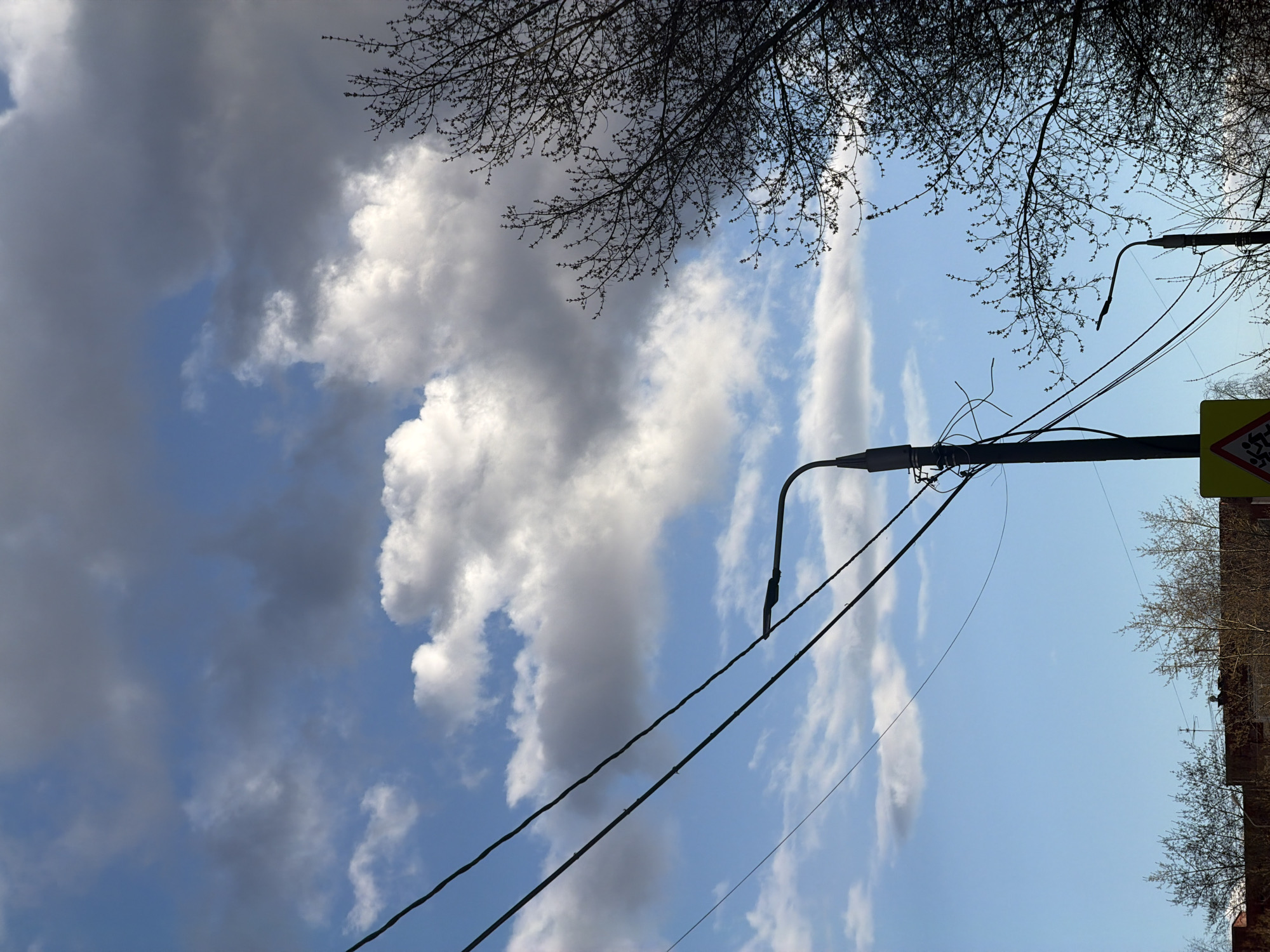 The image, originally rotated, depicts a dynamic urban or suburban sky. The upper left portion of the frame is dominated by large, dark grey clouds, which transition towards the right into bright blue sky interspersed with fluffy white cumulus clouds.
Silhouetted bare tree branches, possibly indicative of late winter or early spring, frame the top and bottom sections of the image. A tall, dark utility pole with an attached streetlight stands prominently on the right side, extending vertically through the frame. Numerous black wires stretch horizontally and diagonally across the sky, connecting to this pole and another partially visible pole on the left.
On the far right, a portion of a yellow, triangular warning sign with a red border is visible. The graphic on the sign depicts two walking figures, suggesting a "children crossing" or "pedestrian crossing" alert. Below this sign, a segment of a brick-colored building is visible, further confirming an urban or built-up environment.
The scene captures a daytime setting, with varied light conditions due to the mix of clear sky and cloud cover. There is no specific activity taking place, and no explicit emotions are conveyed. The photograph provides a snapshot of overhead infrastructure and sky in an urban context, with the specified location being Perm, Russia. No visible text is present other than the graphic on the warning sign.
